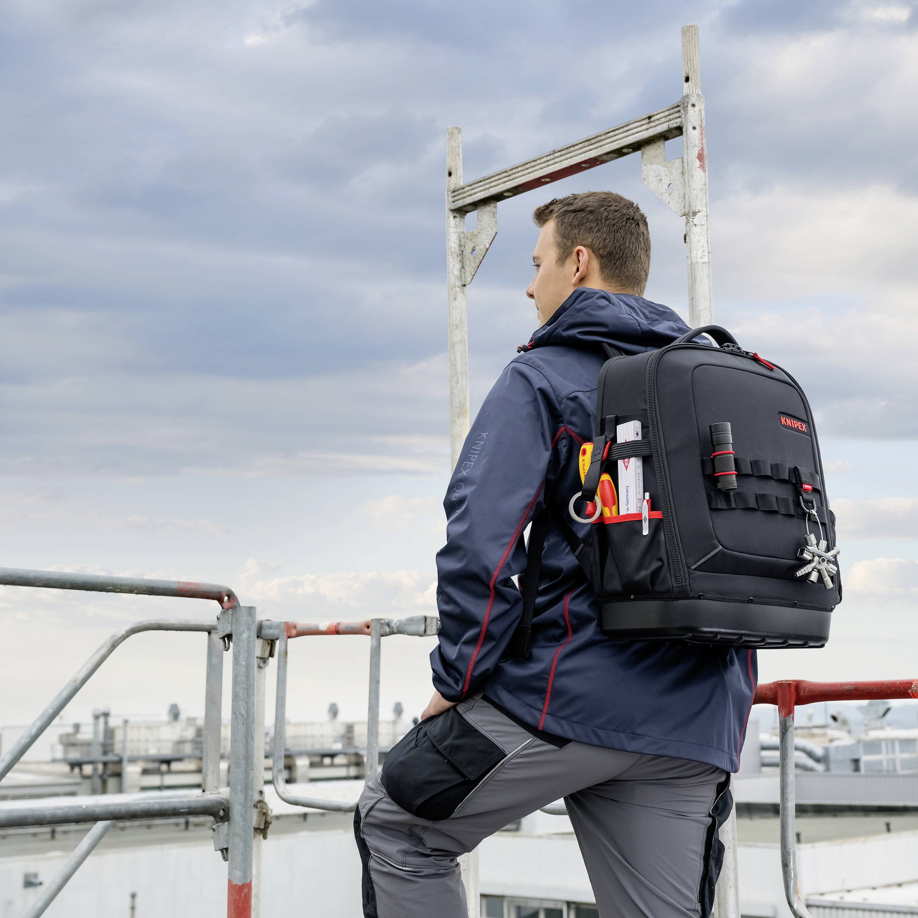 Ein Mann mit einem Werkzeugrucksack steht auf einem Boot oder Steg und blickt über das Wasser unter einem bewölkten Himmel hinaus.