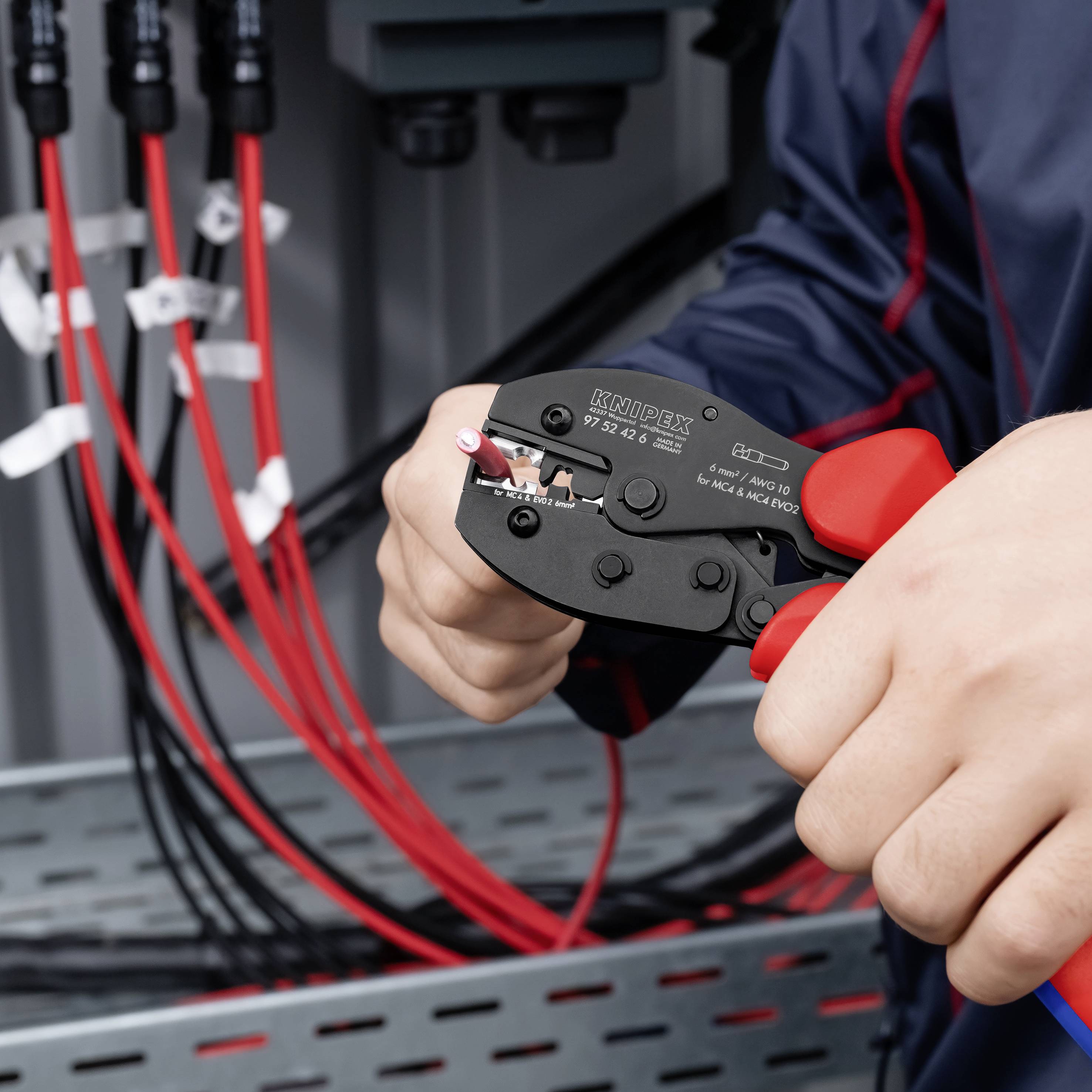Hände, die einen Abisolierzangen verwenden, um ein rotes Kabel in einem Elektroschrank abzuisolieren, wobei andere rote und schwarze Kabel im Hintergrund sichtbar sind.