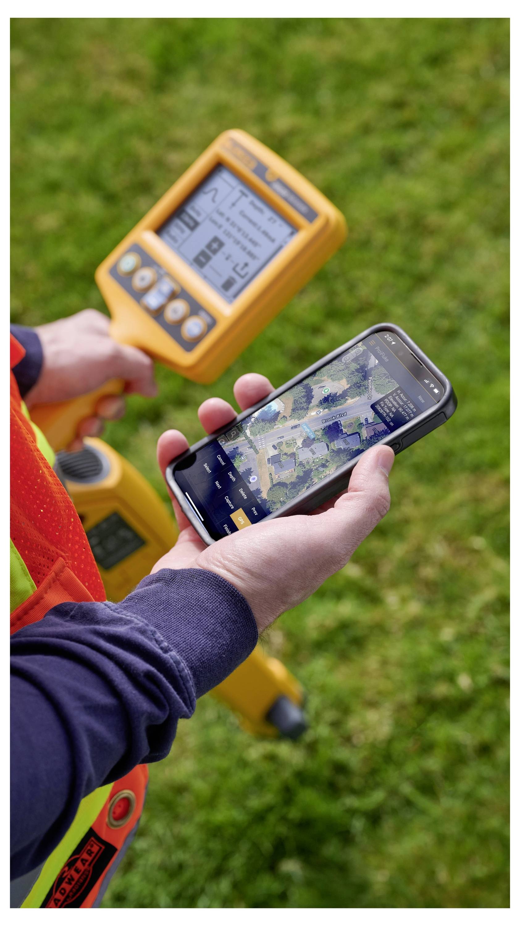 Person in einem Sicherheitsweste nutzt ein Smartphone neben einem tragbaren GPS-Vermessungsgerät im Freien auf einer Grasfläche.