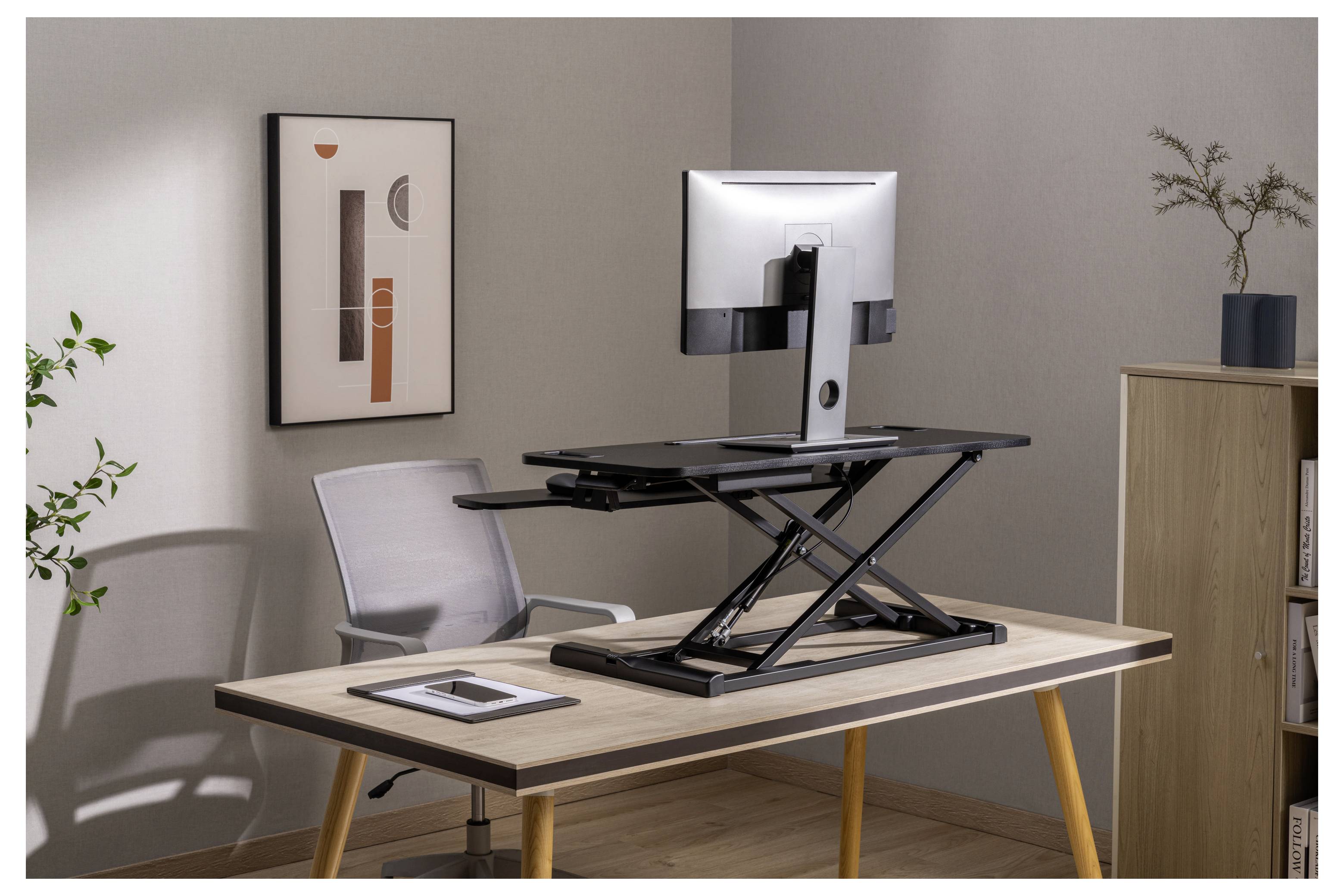 Homeoffice-Schreibtisch-Setup mit einem höhenverstellbaren Sitz-Steh-Schreibtisch, Monitor, Stuhl, Tablet, Wandkunst und Schrank in einem minimalistischen Raum.