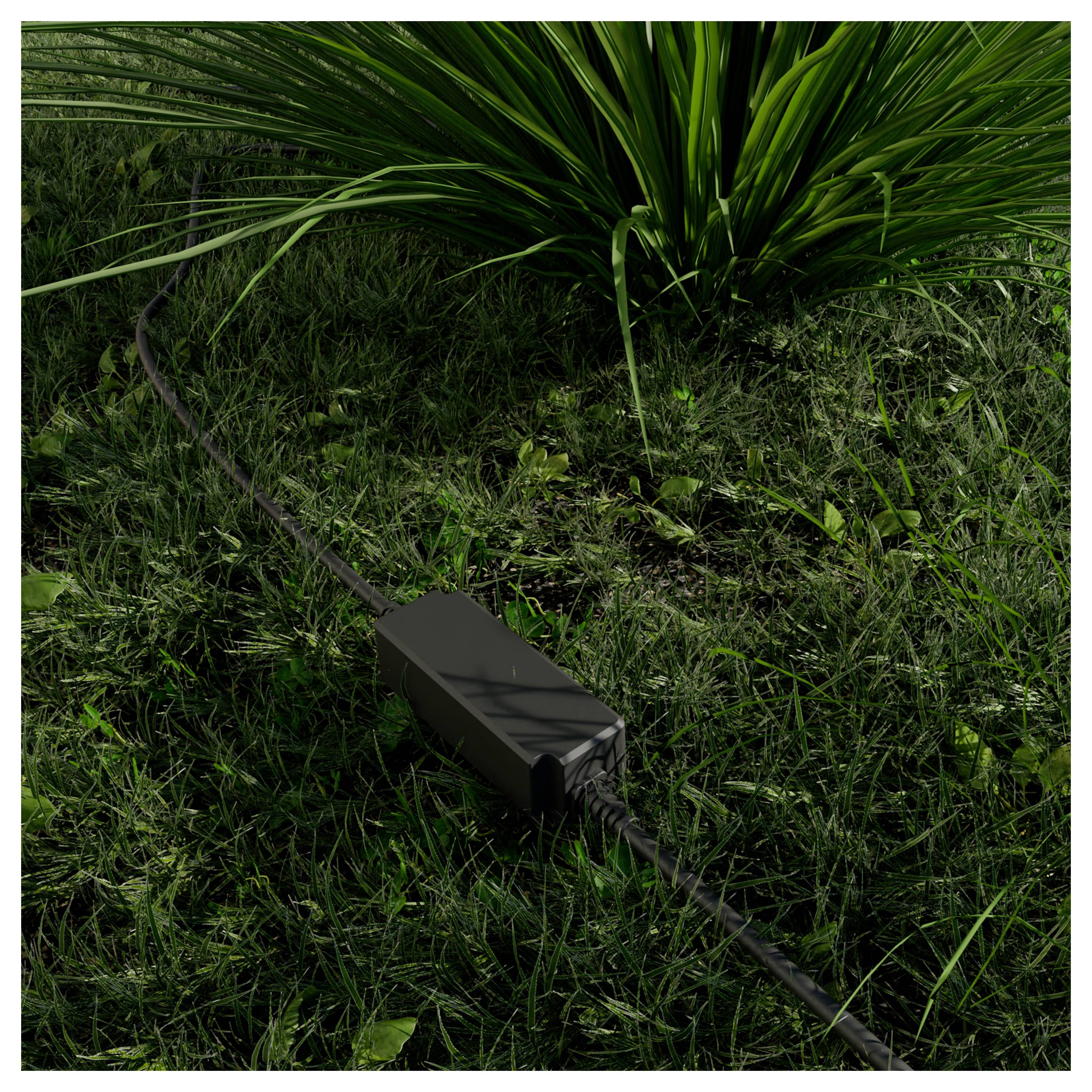 Schwarze Outdoor-Kabelverbindungsdose auf einem Gartenkabel, das durch Gras neben einer stacheligen Pflanze verlegt ist.