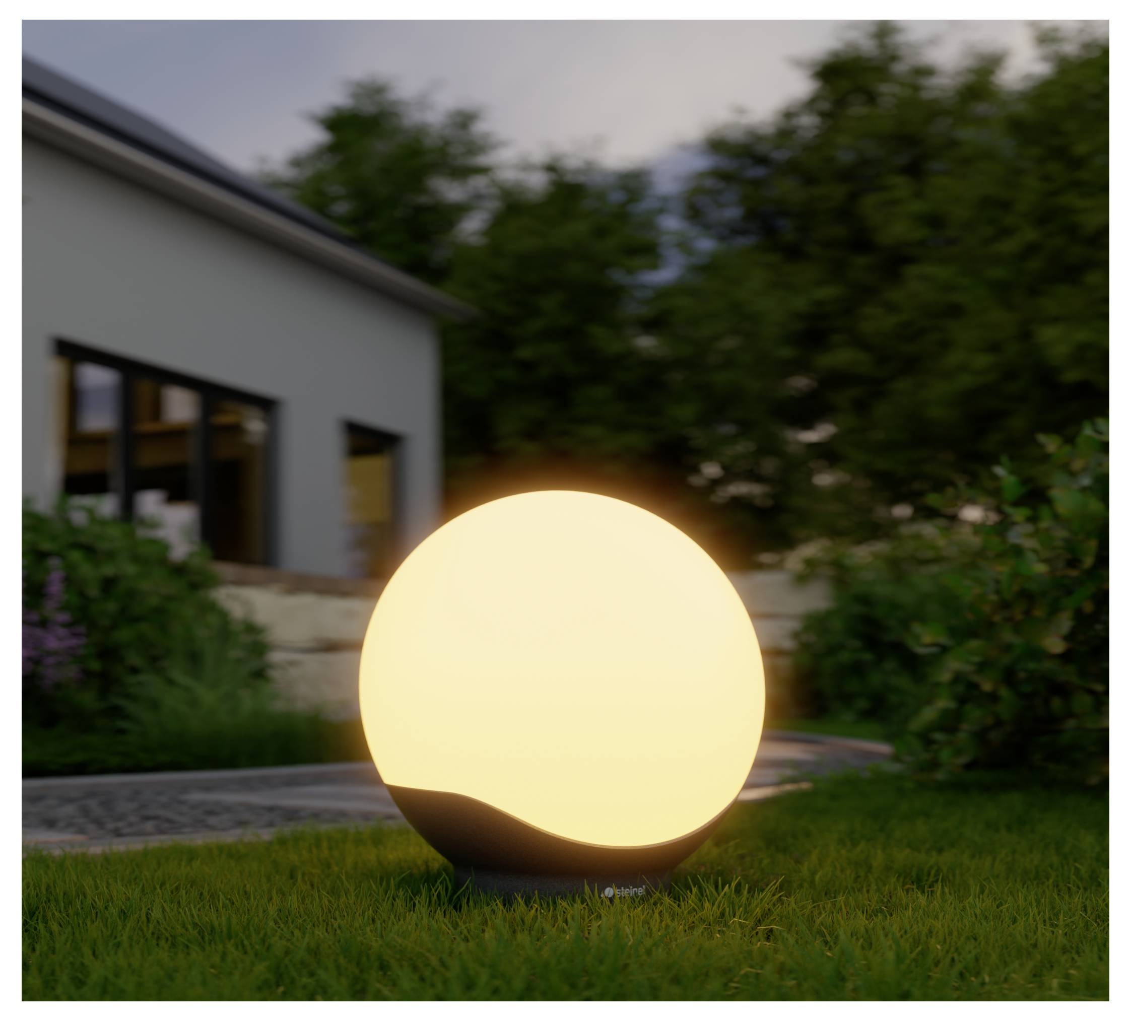Leuchtende kugelförmige Außenleuchte auf Gras in einem Garten in der Dämmerung, neben einem modernen Haus und einer Steinmauer.