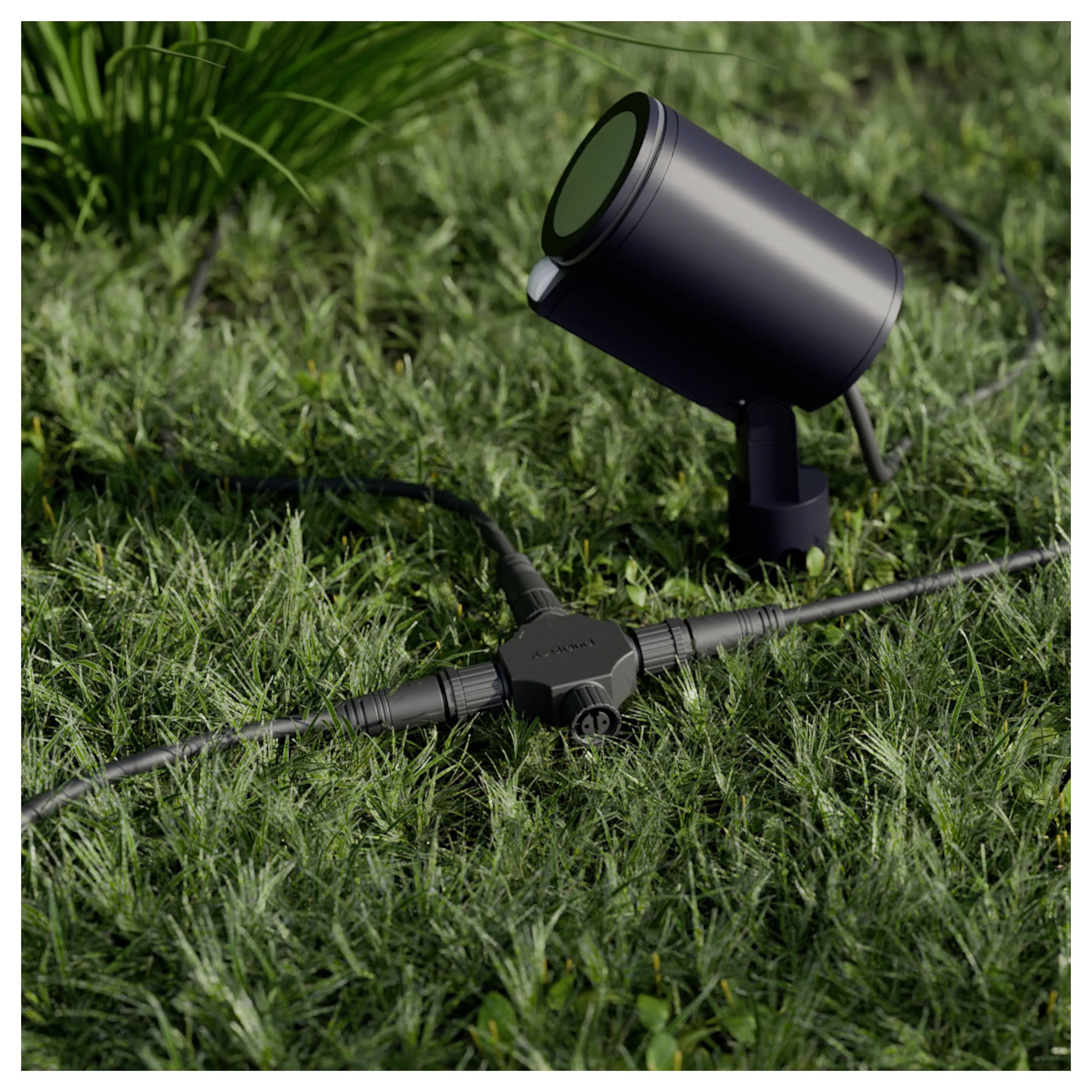 Schwarzer Außenstrahler auf Gras mit angeschlossenem Stromkabel und Anschlussdose, was auf eine Landschaftsbeleuchtungsinstallation hindeutet.