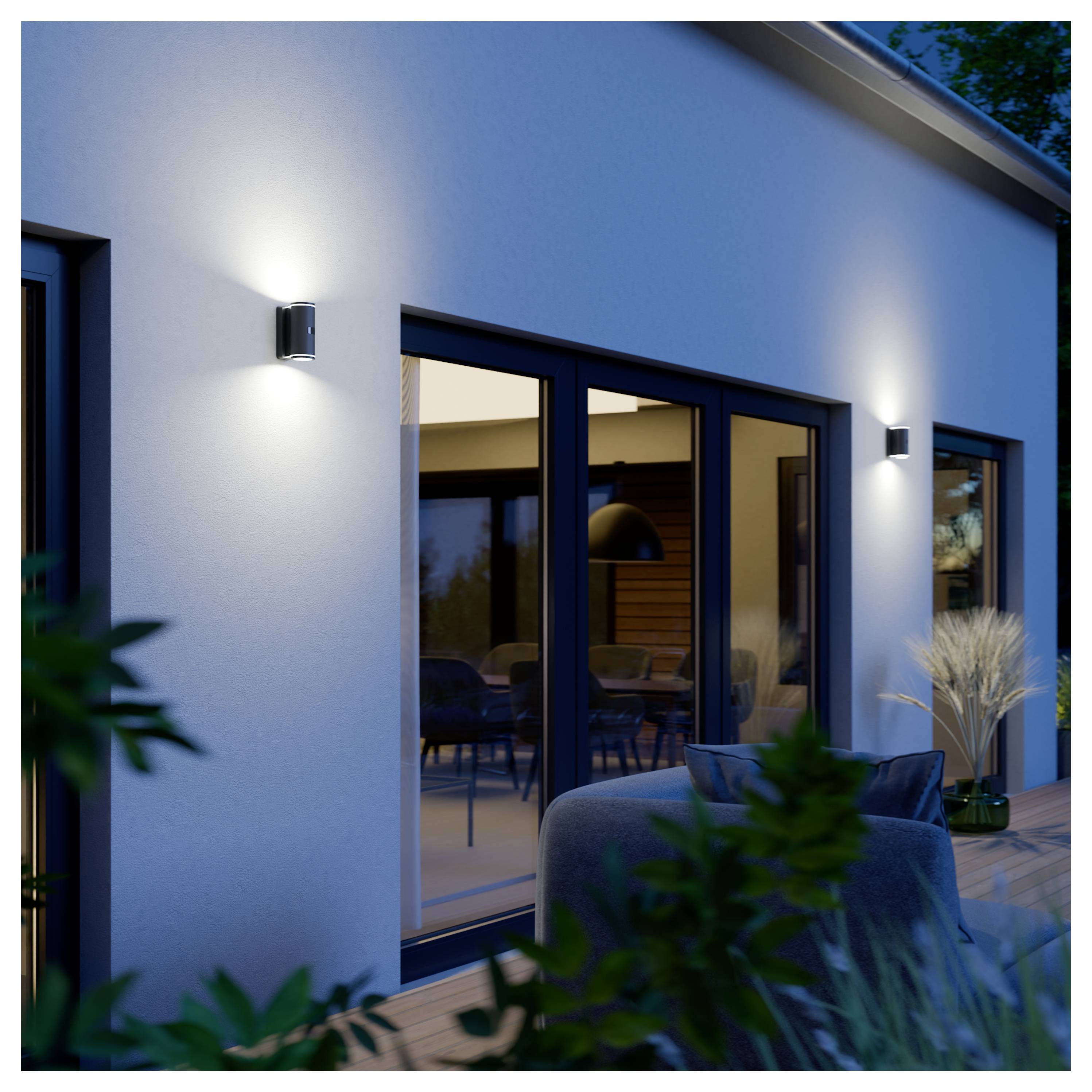 Modernes Hausäußeres in der Abenddämmerung mit beleuchteten Wandleuchten, großen Glasschiebetüren und einer Terrasse mit Sitzmöbeln und Pflanzen.