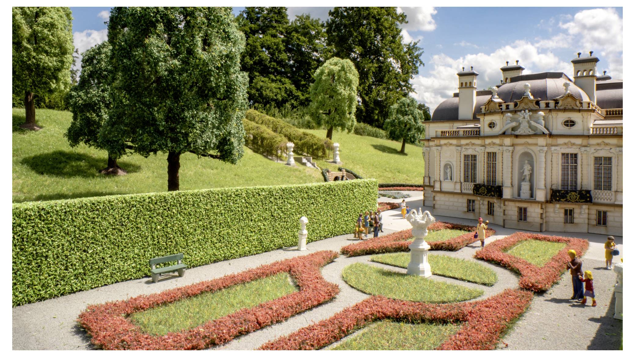 Formaler Garten mit gemusterten Blumenbeeten, Statuen und einem prächtigen historischen Palast neben einer hügeligen Rasenfläche und Bäumen