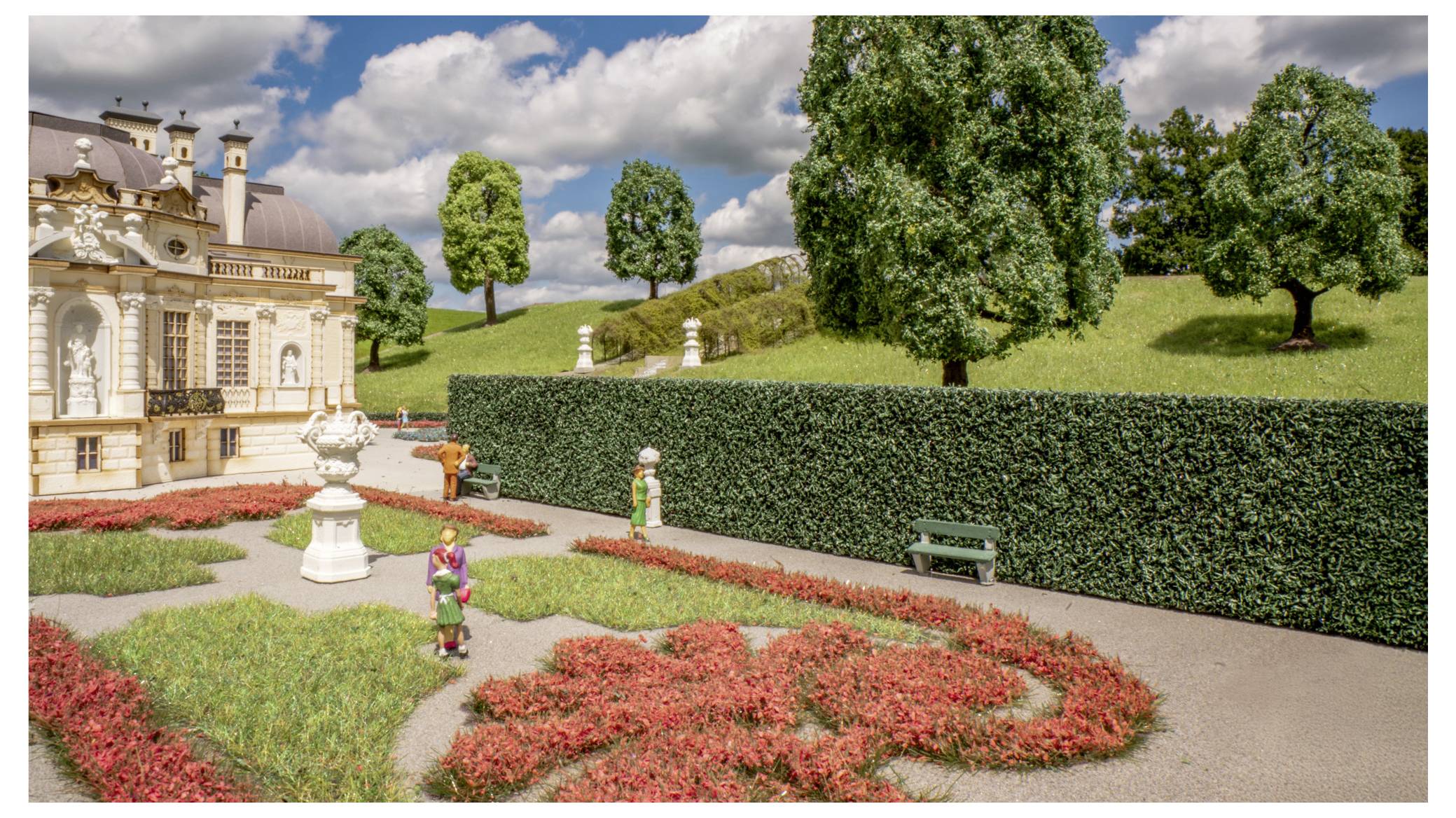 Miniaturformaler Garten neben einem verziertem Gebäude, mit geschnittenen Hecken, Blumenbeeten, Bäumen und kleinen Figuren auf einem Weg unter bewölktem Himmel.