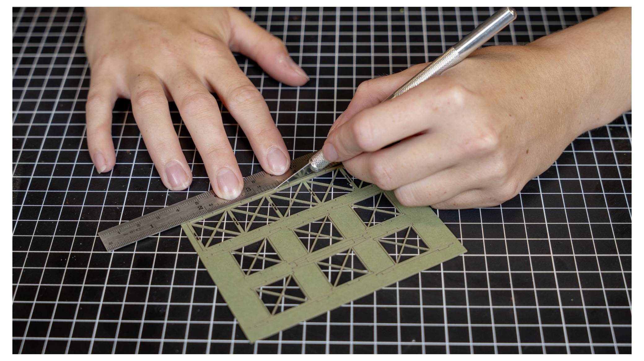 Hände verwenden ein Lineal und ein Handwerksmesser, um ein grünes Gittermuster auf einer Schneidematte zu schneiden.