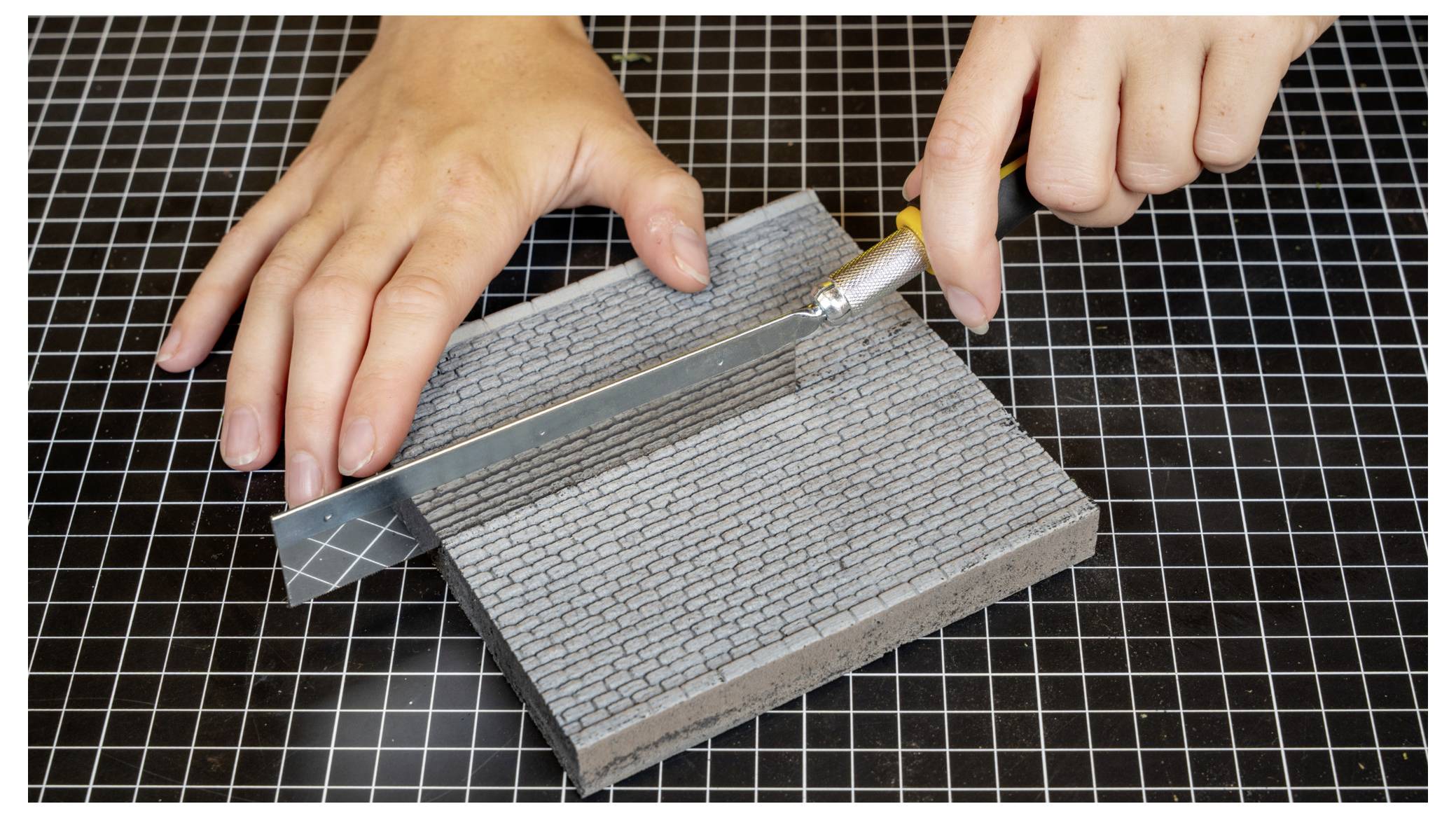 Hände, die ein Bastelmesser verwenden, um einen kleinen strukturierten grauen Modellblock auf einer karierten Schneidematte zu schneiden.