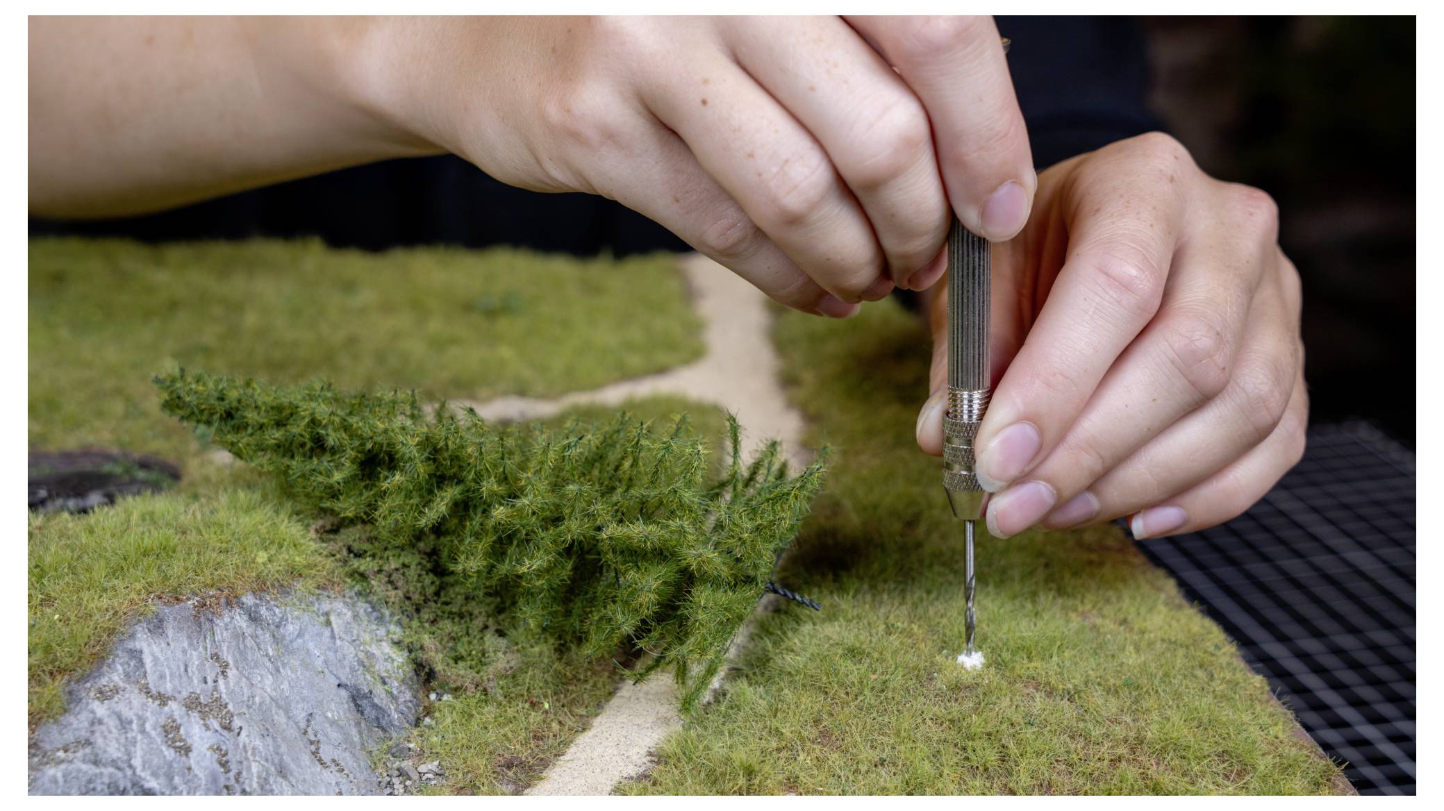 Hände verwenden ein Präzisionswerkzeug, um eine Miniaturmodelllandschaft mit Gras und kleinen Sträuchern zu bepflanzen oder zu reparieren.