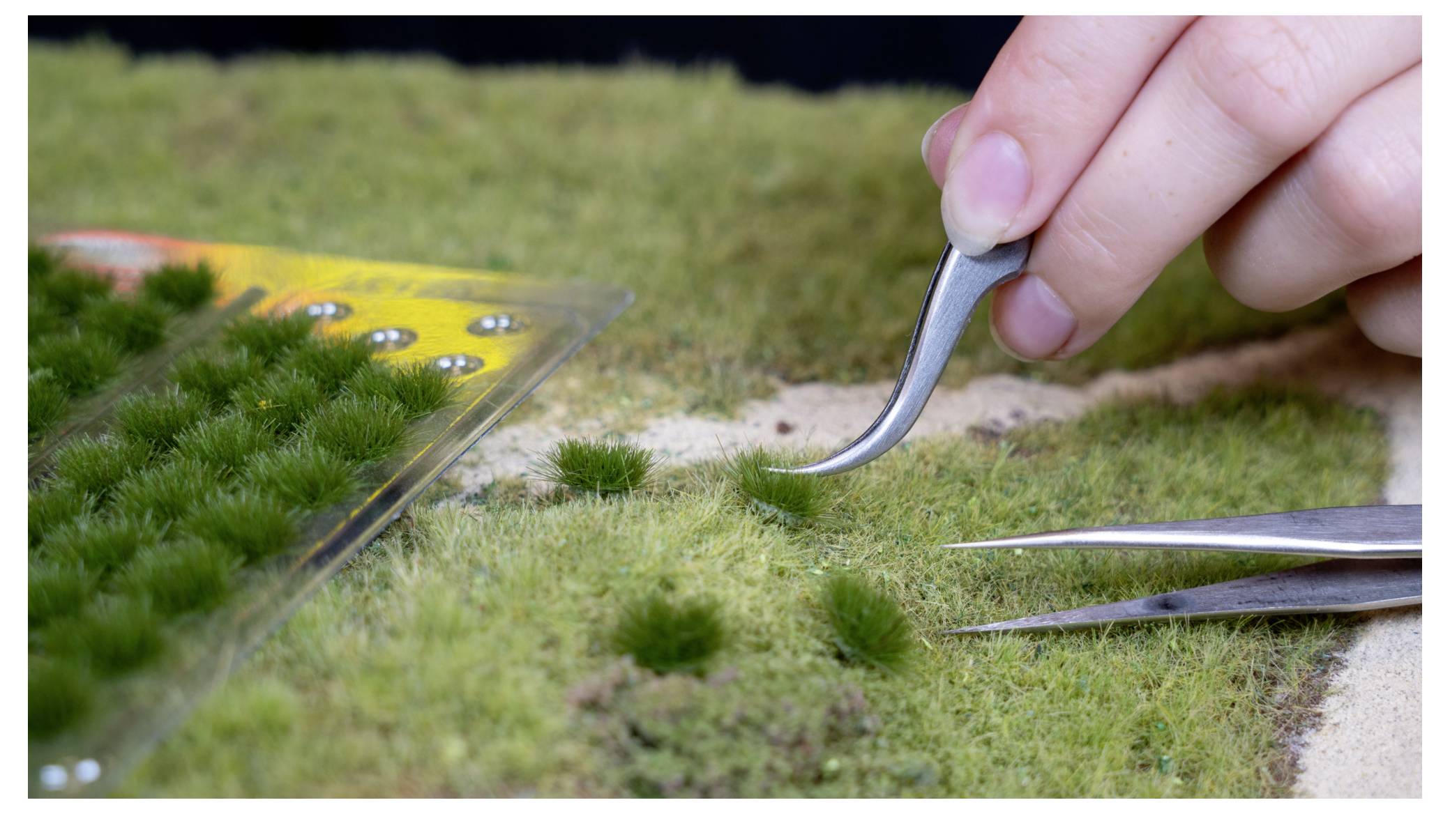 Eine Hand verwendet eine Pinzette, um winzige Geländedetails auf einer Miniaturlandschaft zu platzieren oder anzupassen, wobei sich in der Nähe andere Präzisionswerkzeuge befinden.