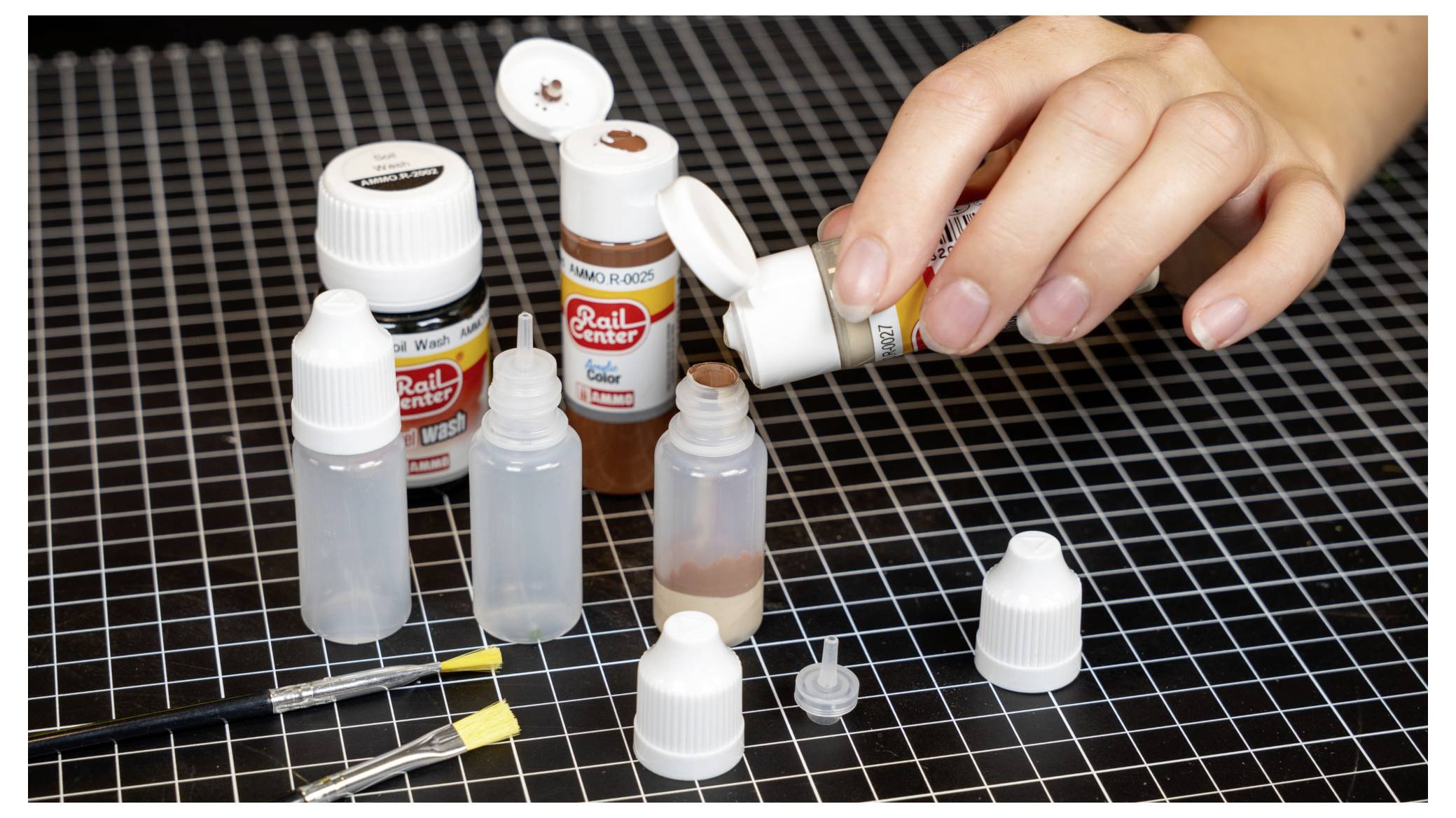 Eine Hand füllt eine kleine Tropfflasche mit Bastelfarbe auf einer karierten Schneidematte, umgeben von mehreren Farbflaschen und Mischungswerkzeugen.