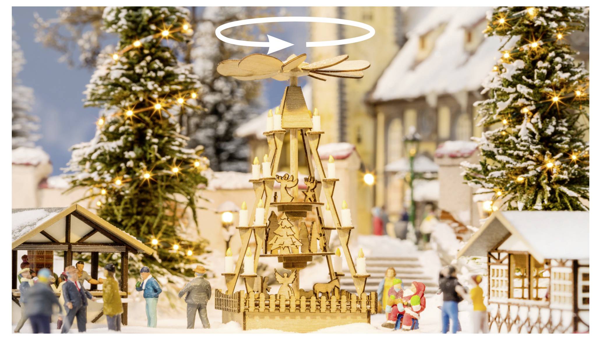 Eine hölzerne Weihnachtspyramide als Tischaufsatz dreht sich über einem verschneiten miniaturhaften Feiertagsdorf mit beleuchteten Bäumen und winzigen Figuren.