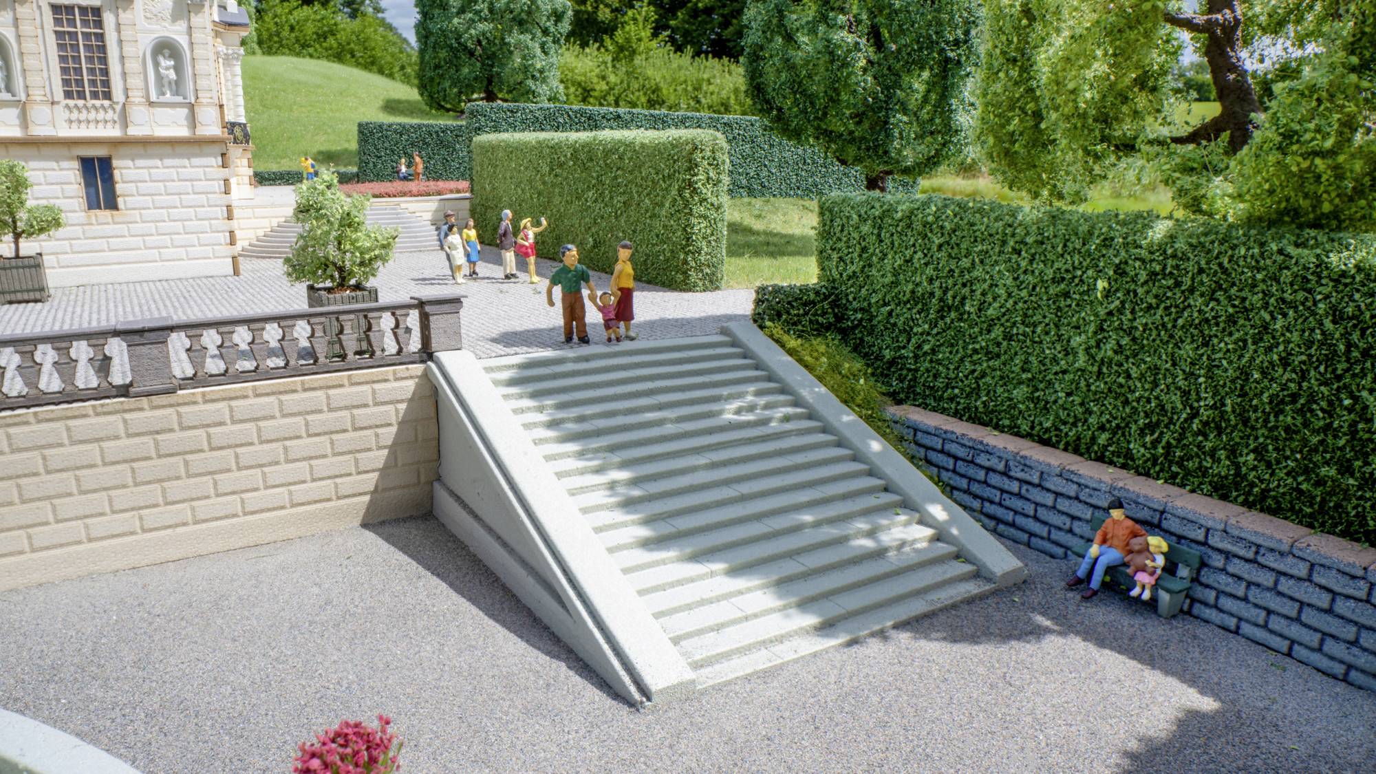 Miniaturansicht einer Außenszene mit einer Rampe mit Stufen, die von einer Terrasse zu einem Kiesweg führt, kleine Figuren in der Nähe und Hecken sowie ein Gebäude im Hintergrund.