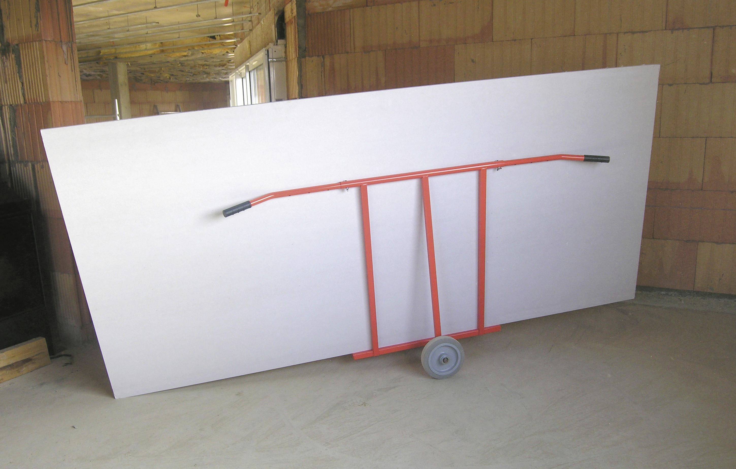 Roter Wagen mit Rädern zum Transport und Heben von Gipskartonplatten, der eine große weiße Platte in einer Werkstatt oder einem Lagerhaus hält.