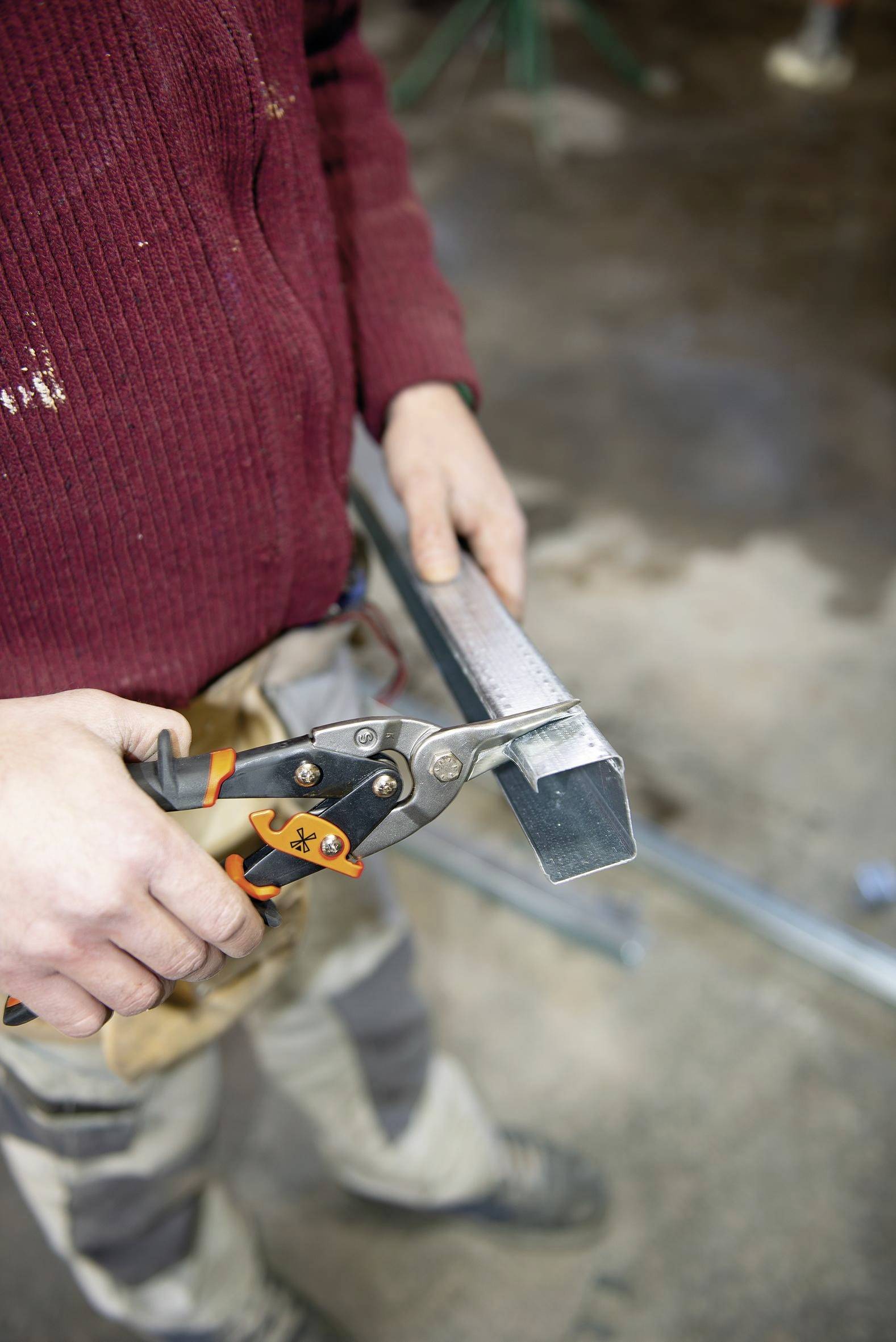 Eine Person in einem roten Pullover benutzt eine Blechschere, um einen Metallstreifen in einer Werkstatt zu schneiden.
