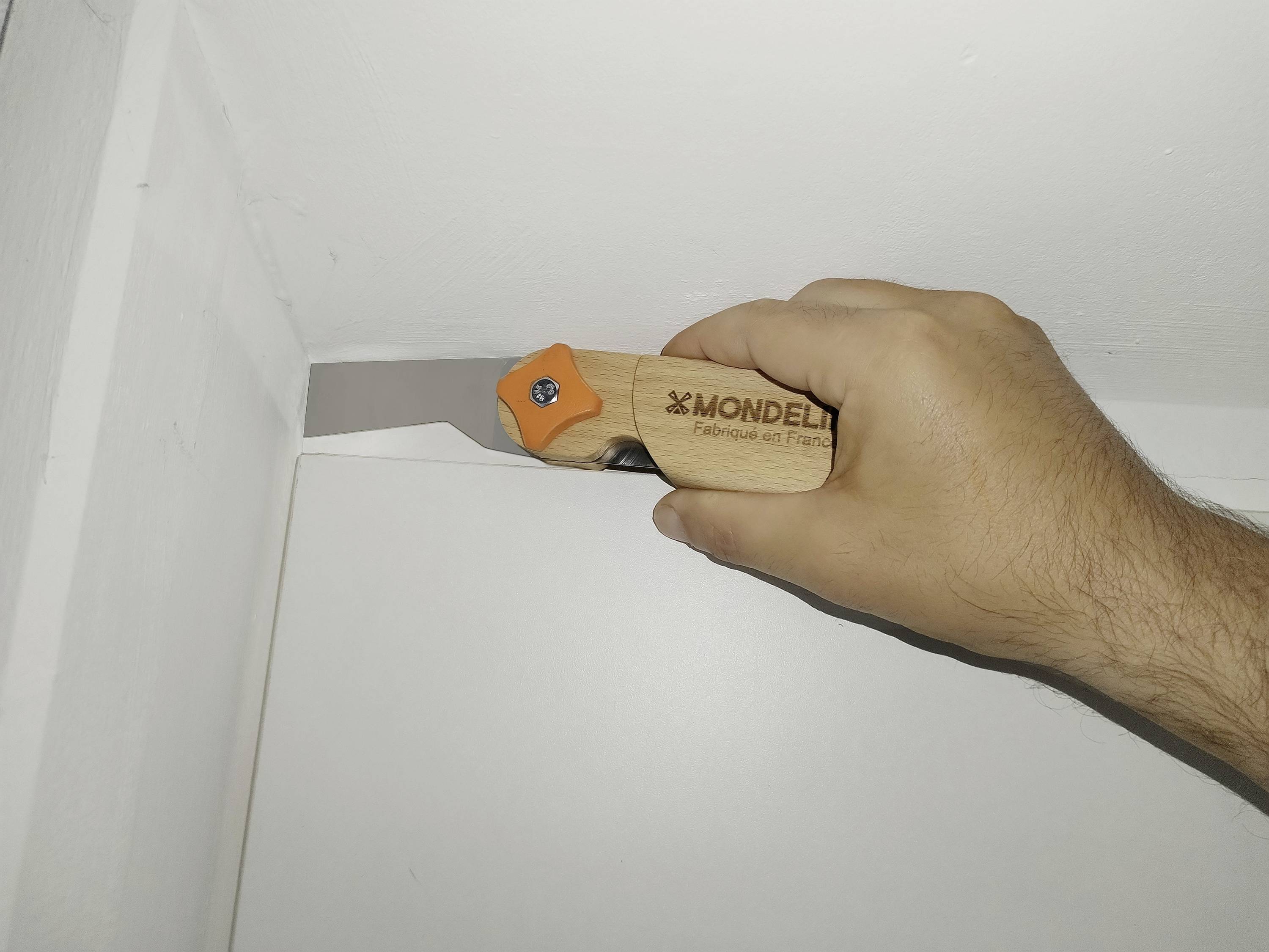 Eine Hand benutzt ein kleines Universalmesser, um entlang der oberen Kante einer weißen Schachtel oder eines Paneels an einer Wand zu schneiden.