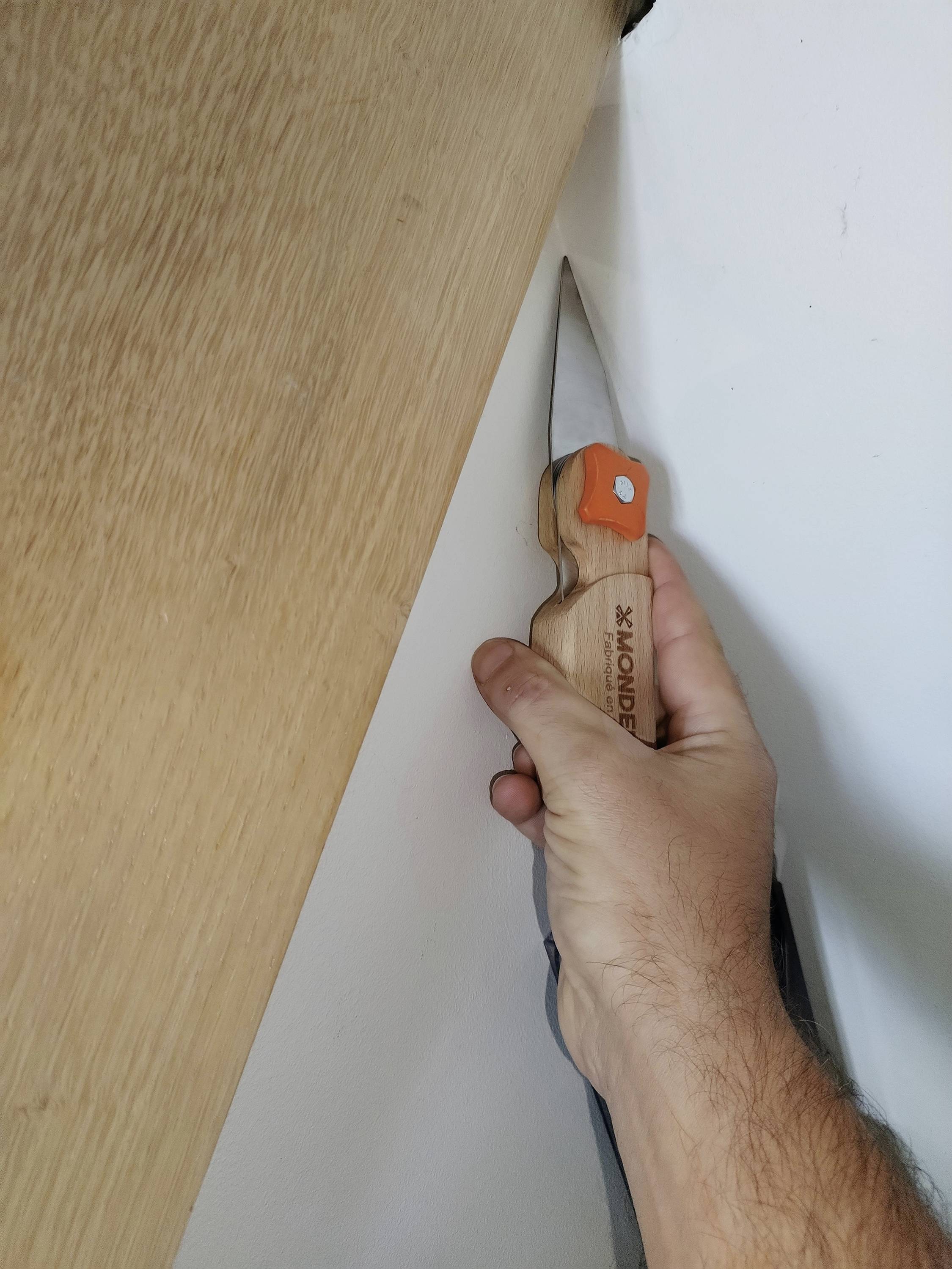 Eine Hand, die ein Universalmesser hält und entlang der Kante einer Holzplatte gegen eine weiße Wand schneidet.
