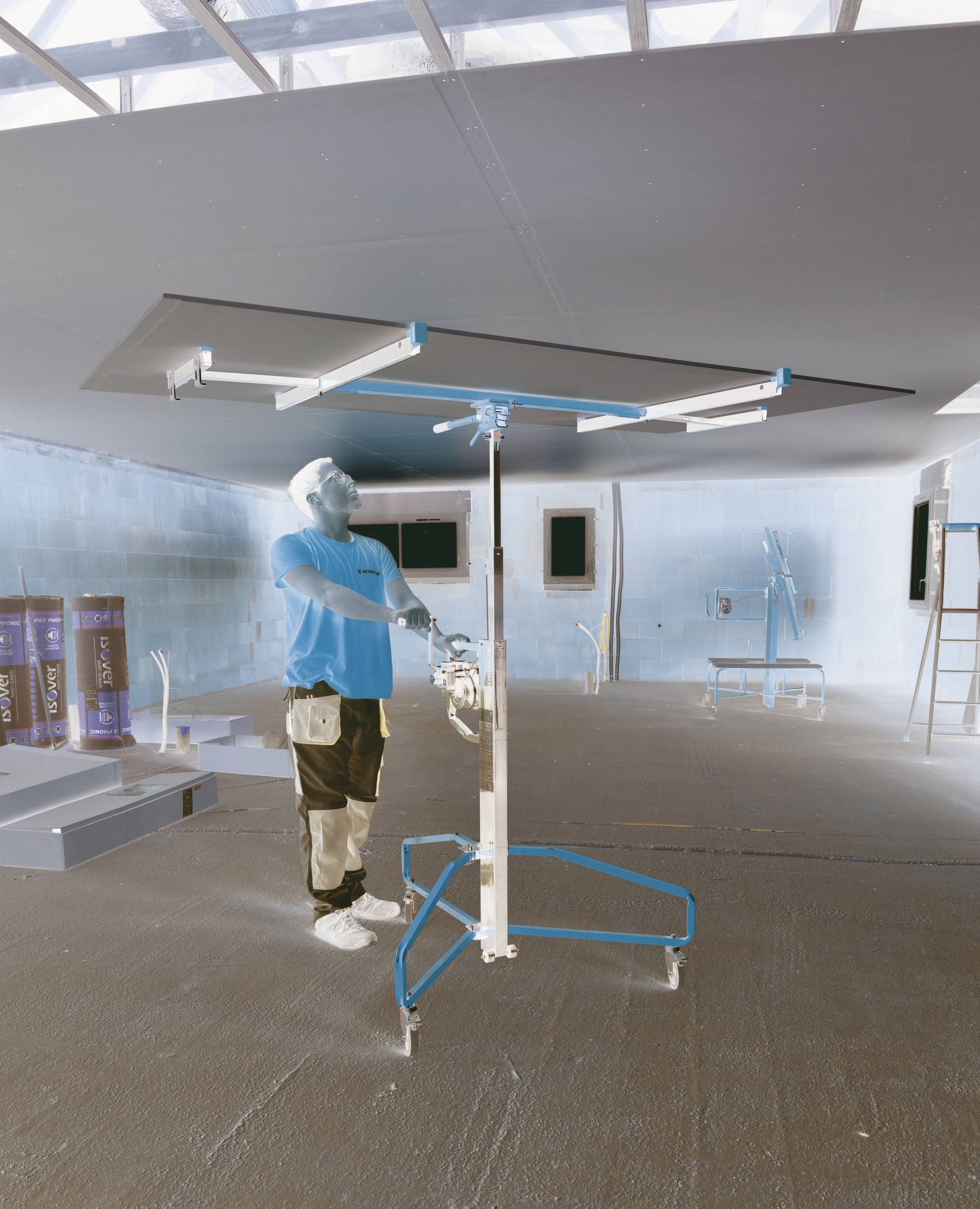 Ein Arbeiter sprüht oder streicht eine große Innenendecke mit Hilfe eines fahrbaren Stützgerüsts in einem größtenteils leeren Raum, der sich im Bau oder in der Renovierung befindet.