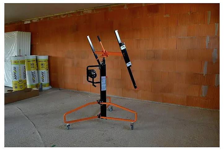 Orange-schwarzer Trockenbaulift auf Rollen in einem unfertigen Raum mit sichtbaren Ziegelmauern und Baumaterialien.