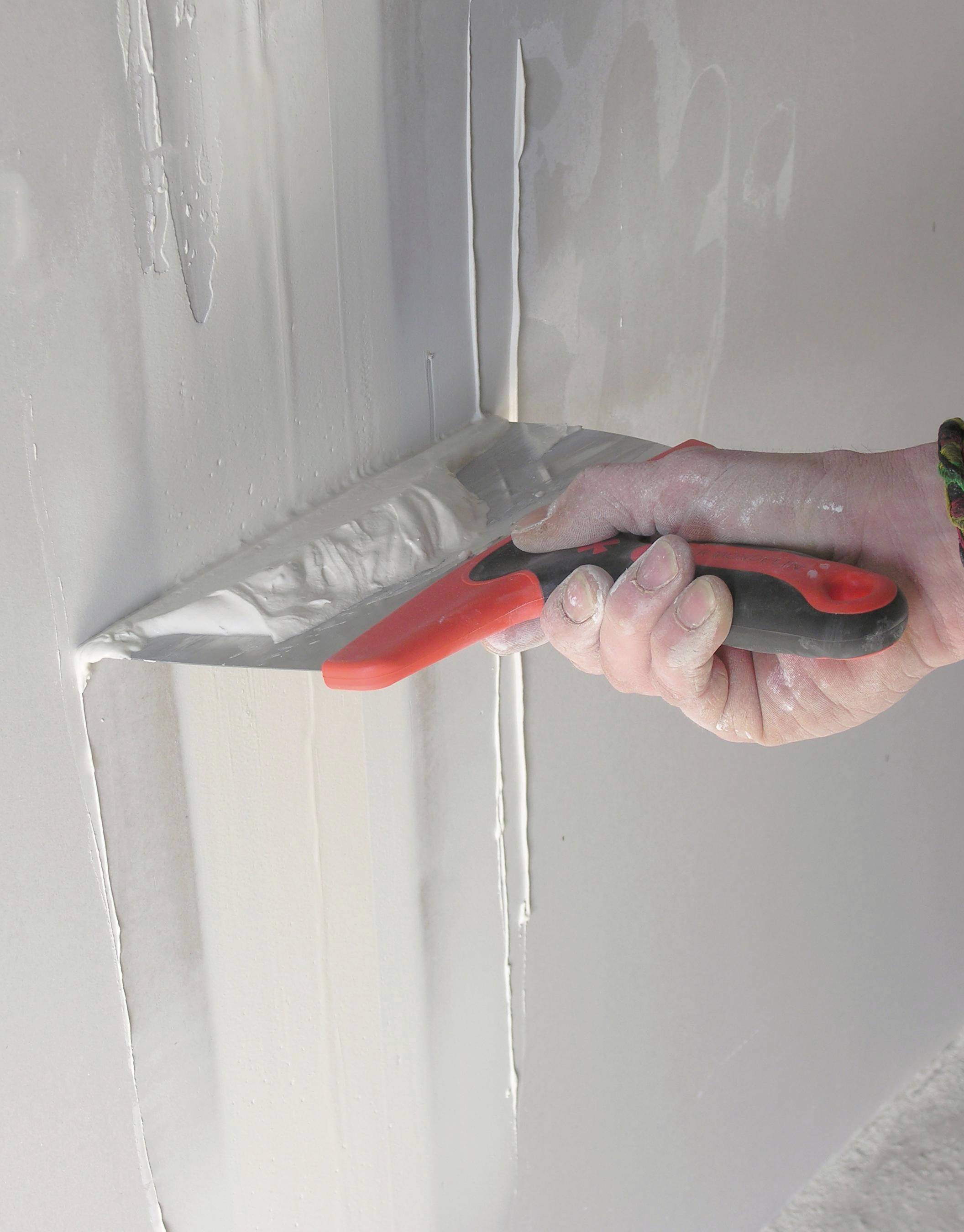 Hand, die einen rot-schwarzen Spachtel verwendet, um graue Fugenmasse in einer Wandecke während der Trockenbaufinish zu glätten.