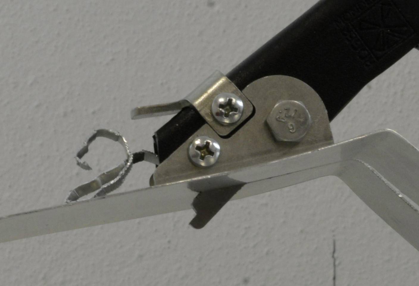 Nahaufnahme einer Metallhalterung und einem schwarzen Riemen, die mit Schrauben auf einer wandmontierten Oberfläche befestigt sind, mit einem kleinen beschädigten Materialstreifen in der Nähe.