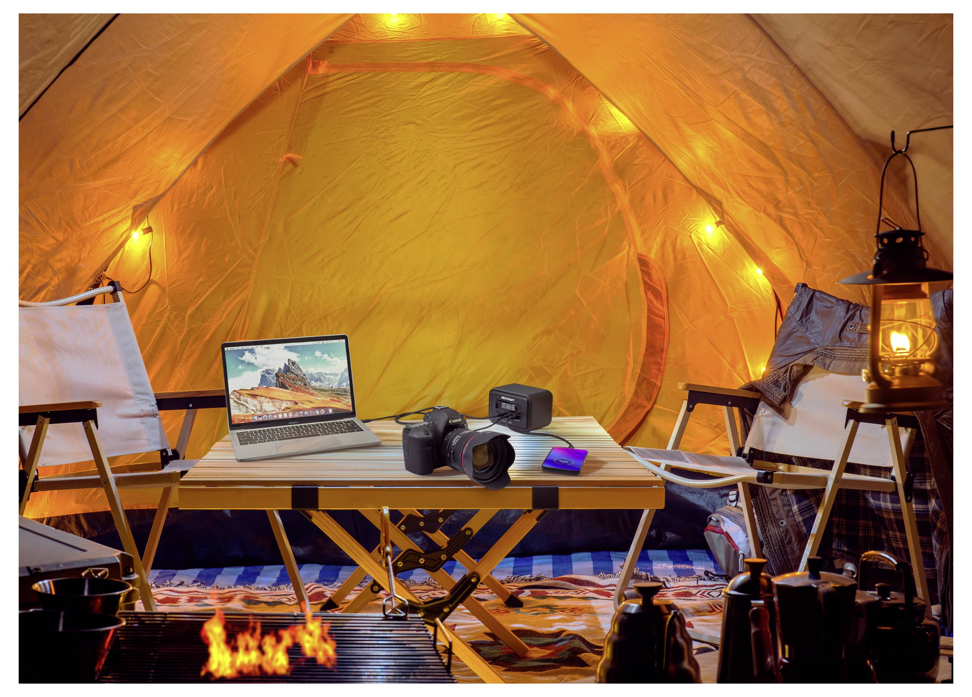 Warmly beleuchtetes Campingzeltinneres mit einem Laptop, einer Kamera, einem Rucksack, Stühlen, Laternen und einem kleinen Feuer vor einem Tischaufbau.