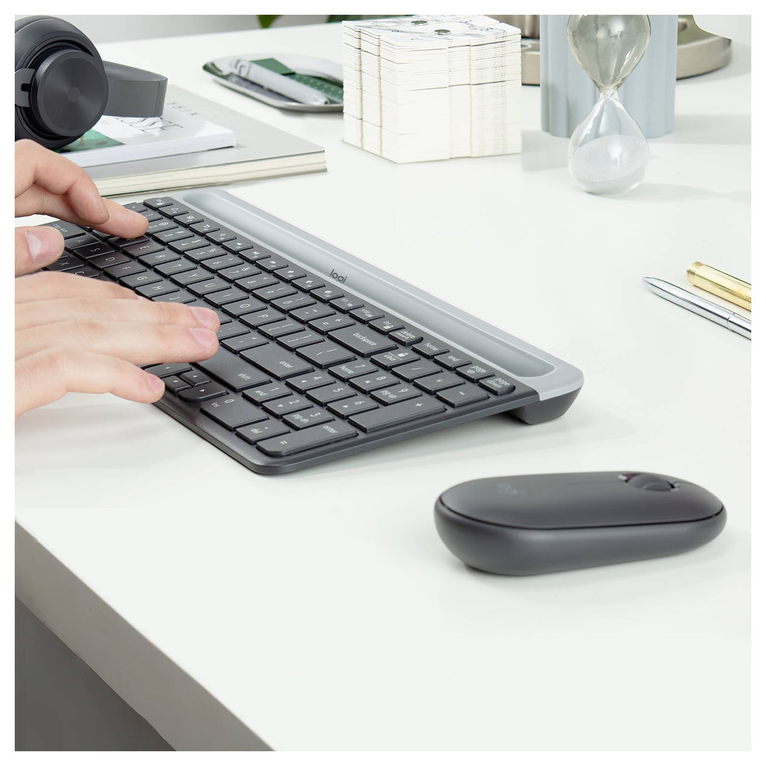 Hände tippen auf einer schwarzen kabellosen Tastatur an einem weißen Schreibtisch mit Maus, Kopfhörern, Notizbuch, Stiften und Sanduhr.