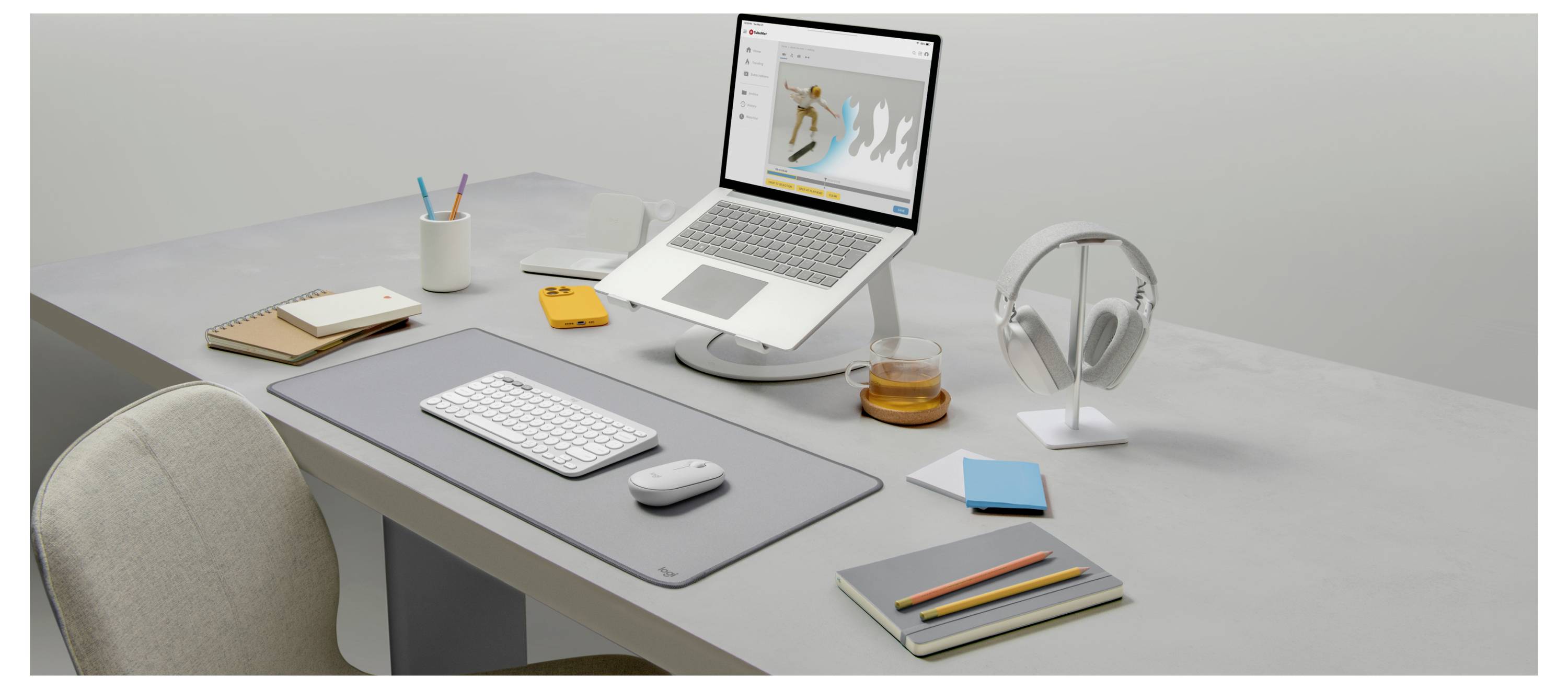 Minimaler Desktop-Arbeitsplatz mit Laptop, Tastatur, Maus, Notizbuch, Bleistiften, Kopfhörern und einem Getränk auf einem grauen Schreibtisch.