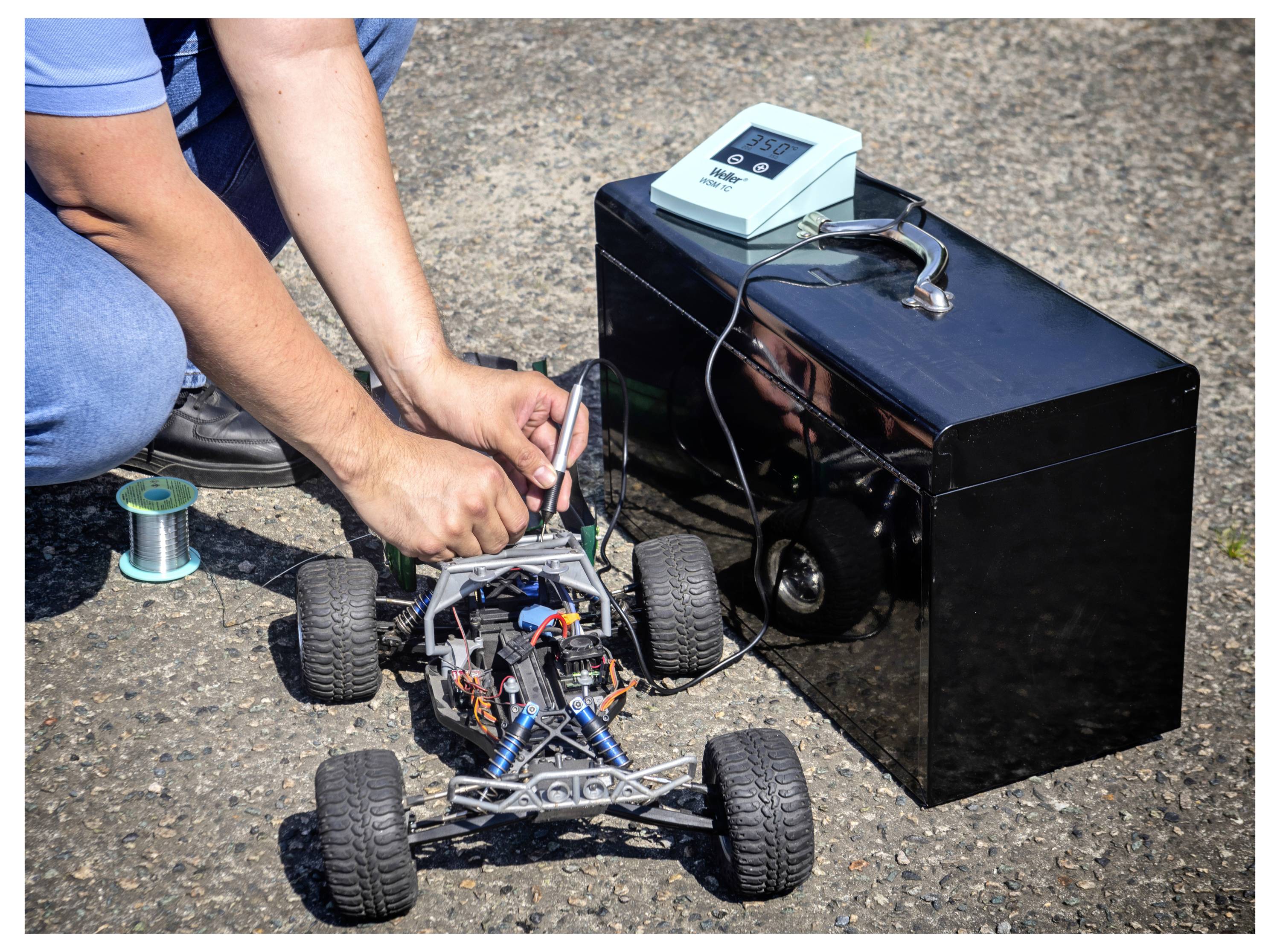 Person, die ein kleines ferngesteuertes Auto auf dem Pflaster neben einem schwarzen Gerät mit einem Messinstrument auf der Oberseite anpasst.