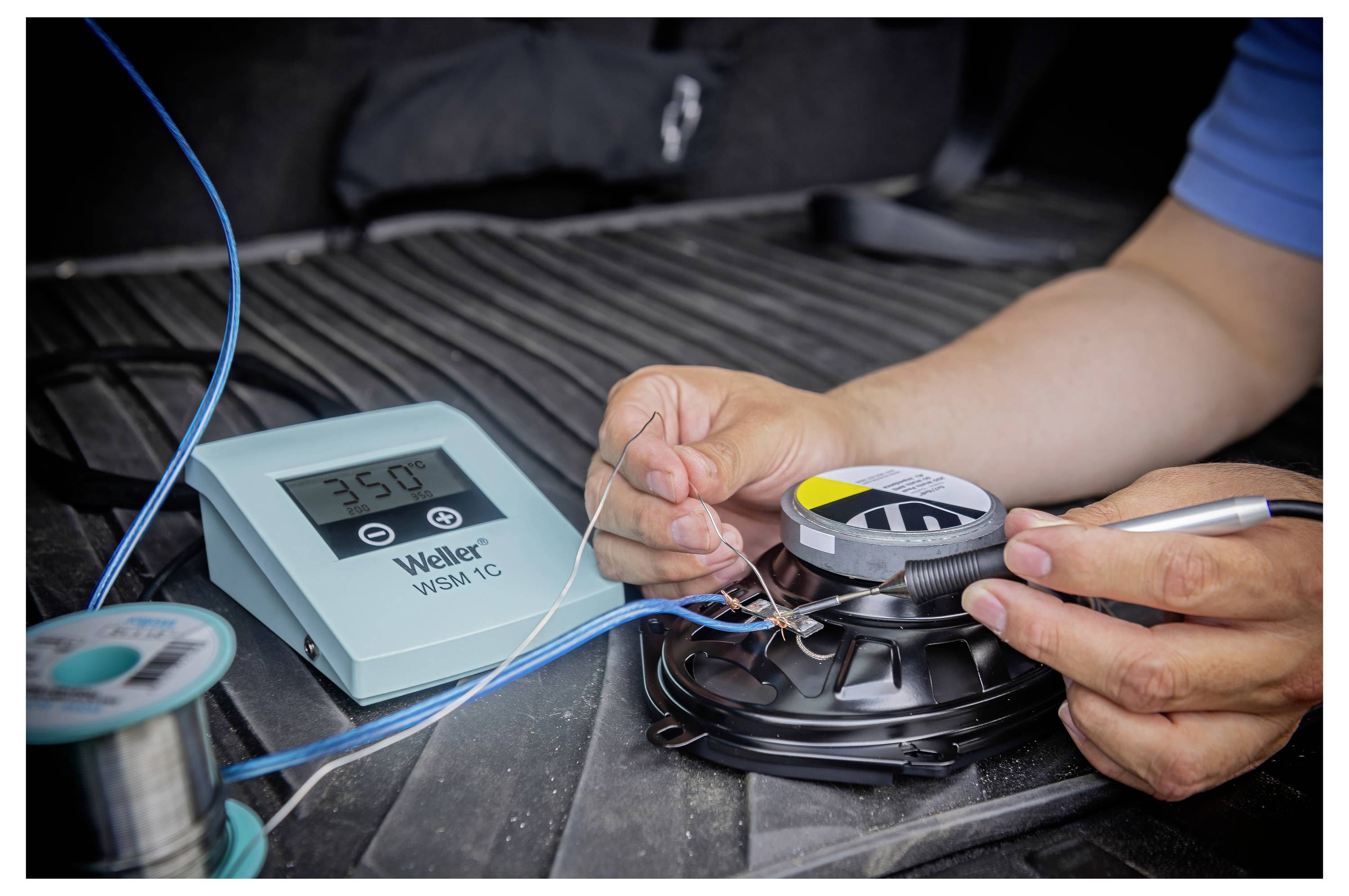 Hände, die mit einem Lötkolben Drähte an einer Lautsprecherkomponente im Kofferraum eines Autos verbinden, mit einer Weller-Lötstation in der Nähe.