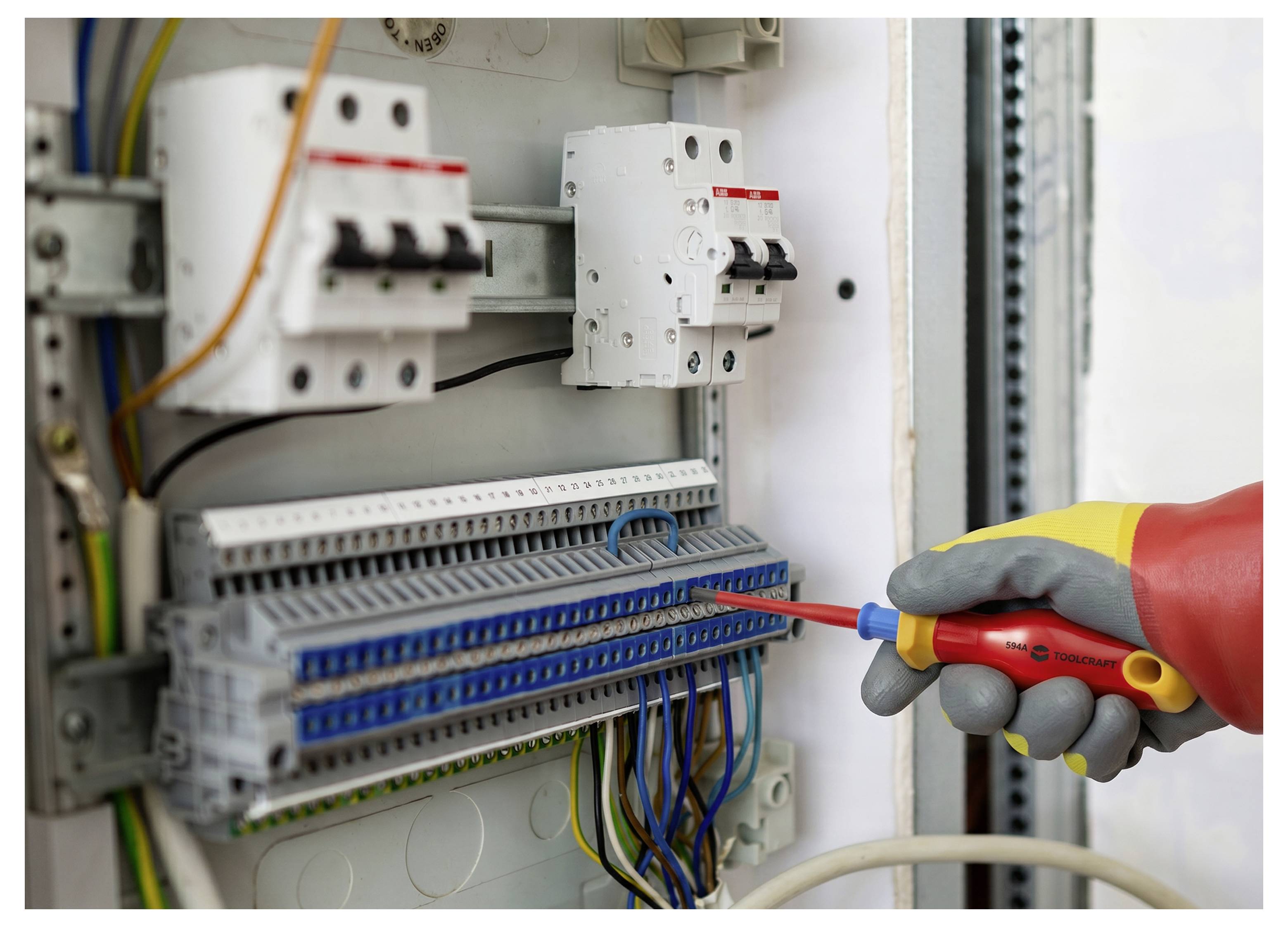 Mit Handschuhen bekleidete Hand verwendet einen isolierten Schraubendreher, um die Verdrahtung in einem elektrischen Schaltschrank mit Leistungsschaltern und Klemmenleisten einzustellen.