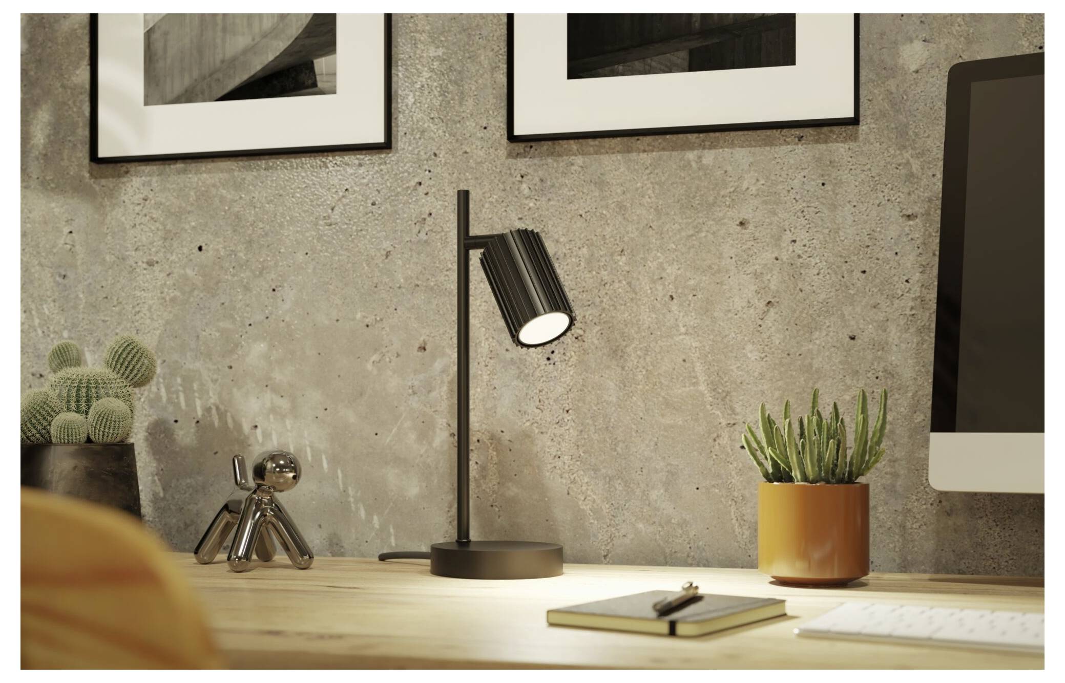 Modernes Schreibtischset-up mit leuchtender schwarzer Tischlampe, Topfpflanze, Notizbuch und Computermonitor vor einer Betonwand mit gerahmter Kunst.