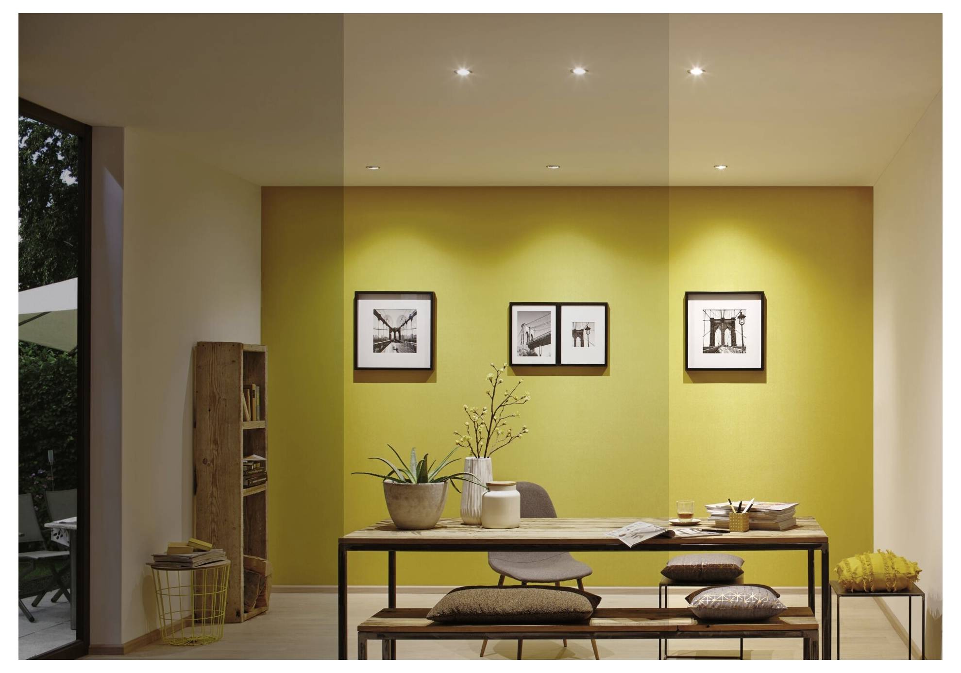 Modernes Esszimmer mit einem Holztisch, Sitzbänken und gerahmten Wandkunstwerken an einer leuchtend gelben Akzentwand, beleuchtet durch eingelassene Deckenleuchten.