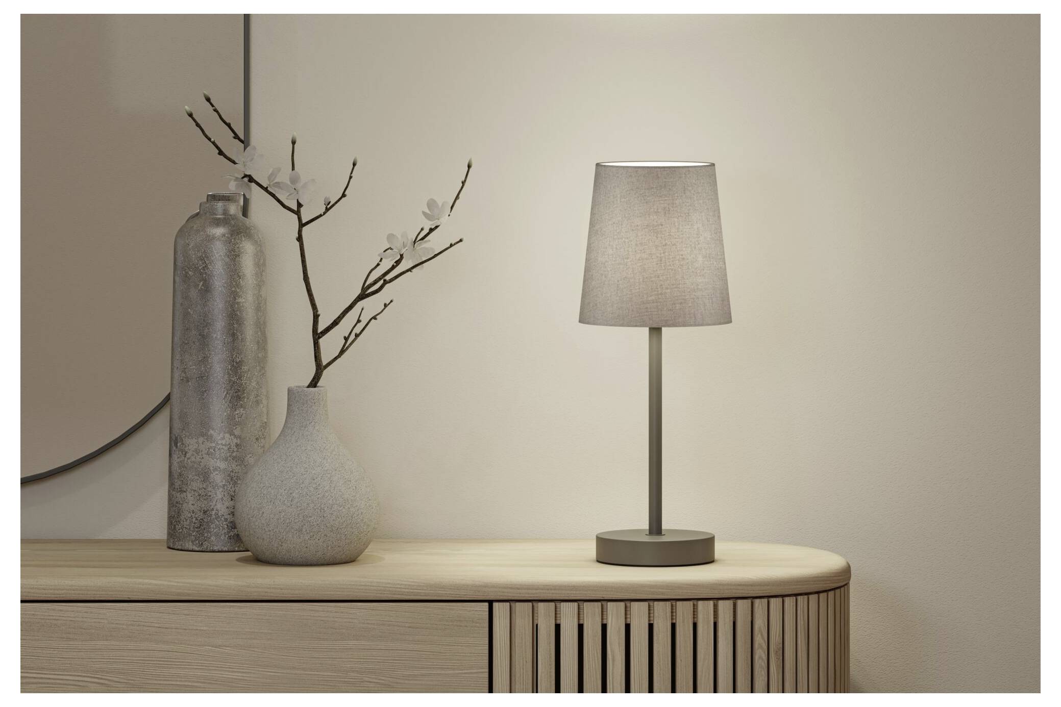 Minimale Innenraumszene mit einer leuchtenden Tischlampe auf einem Holzschrank neben einer Vase mit Zweigen und einem hohen dekorativen Zylinder.