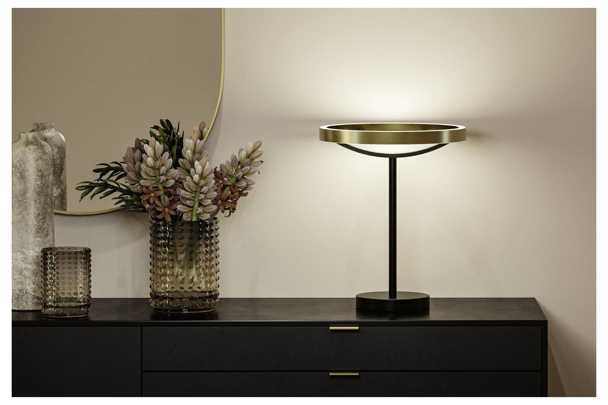 Moderne schwarze Tischlampe mit leuchtendem Ringschirm auf einer dunklen Kommode neben einer Glasvase mit getrockneten Blumen und einem Kerzenhalter.