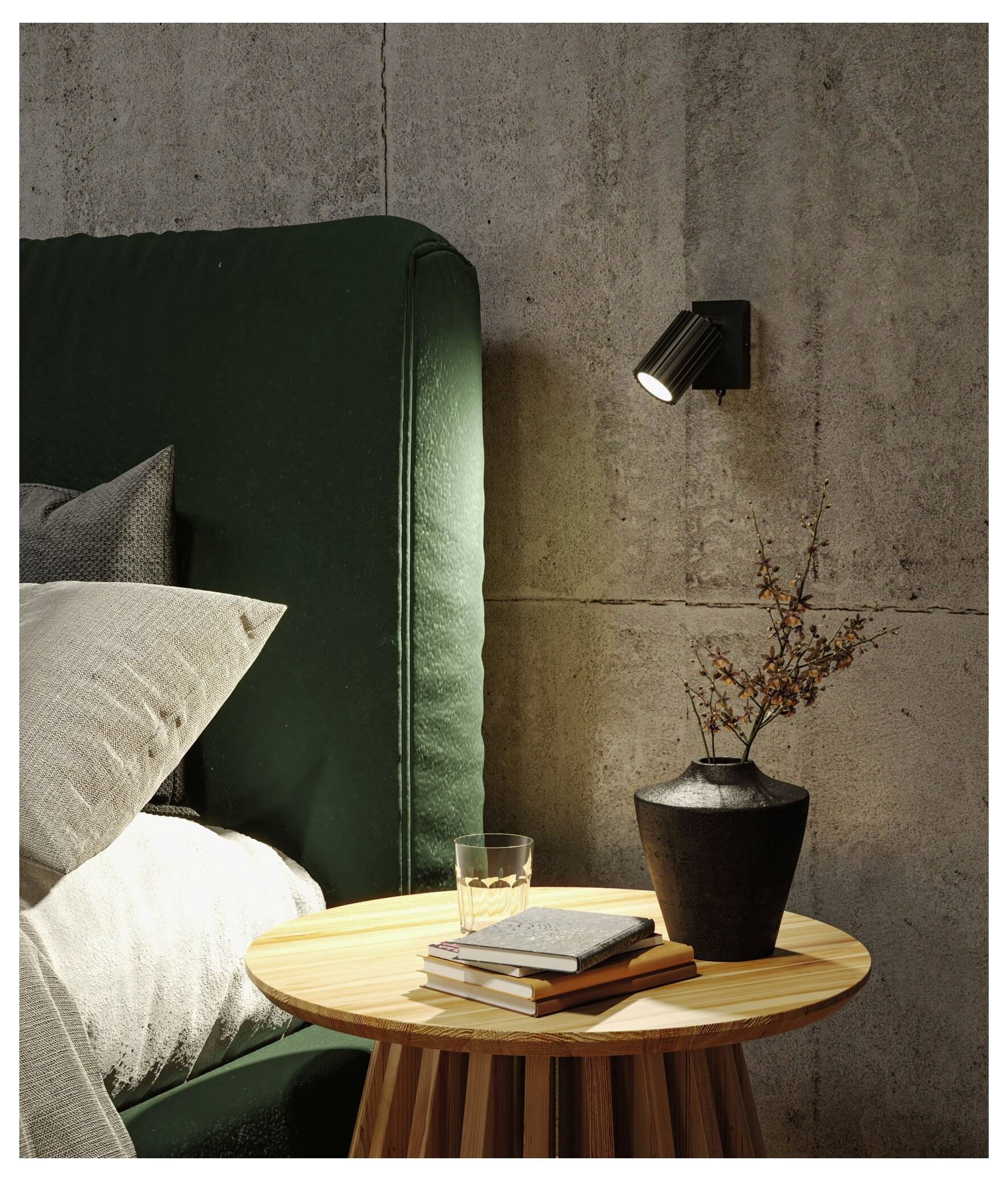 Eine moderne Schlafzimmerecke mit einem grün gepolsterten Bett, rundem Holznachttisch, Büchern, Glas, Vase mit getrockneten Ästen und Wandstrahler.