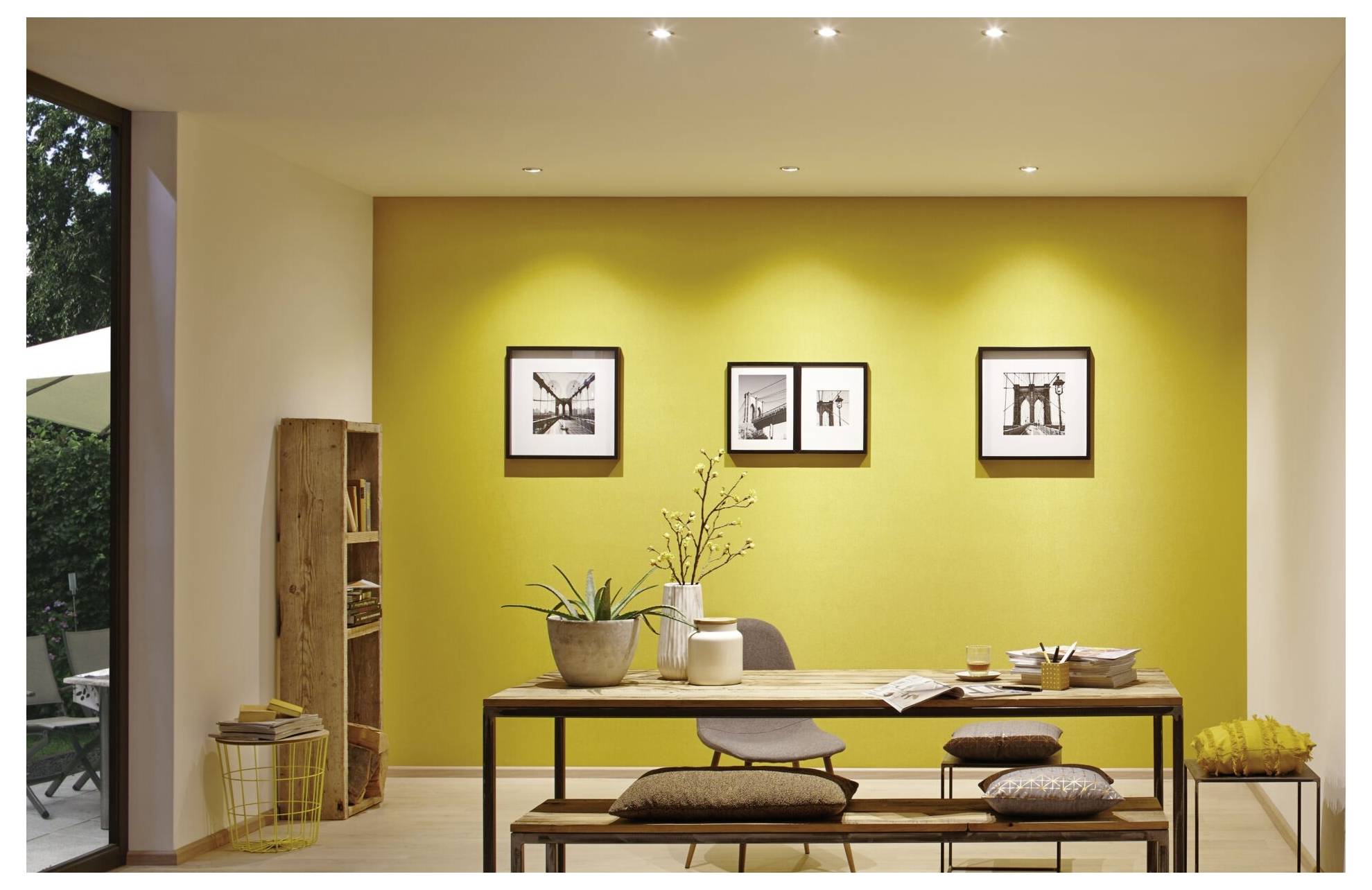 Modernes Esszimmer mit gelber Akzentwand, rahmendem Kunstwerk, Holztisch, Bänken, Pflanzen und sanfter Deckenbeleuchtung.