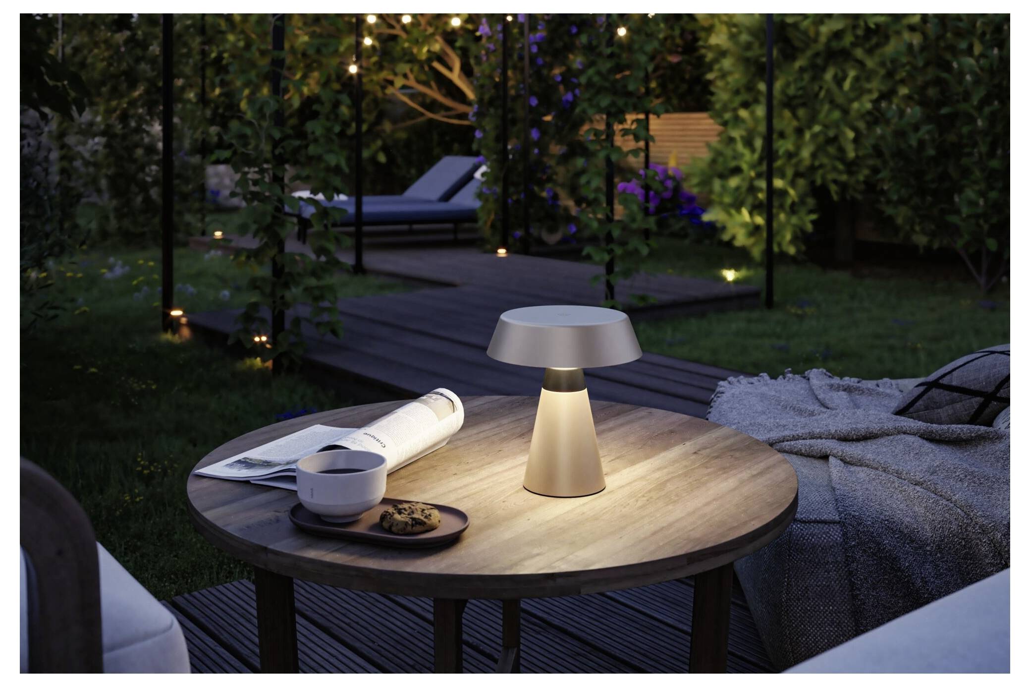 Tragbare Tischleuchte, die einen Außenpatio-Tisch neben einer zusammengerollten Zeitschrift, einer Schüssel und einer Decke in einem Garten in der Dämmerung beleuchtet.