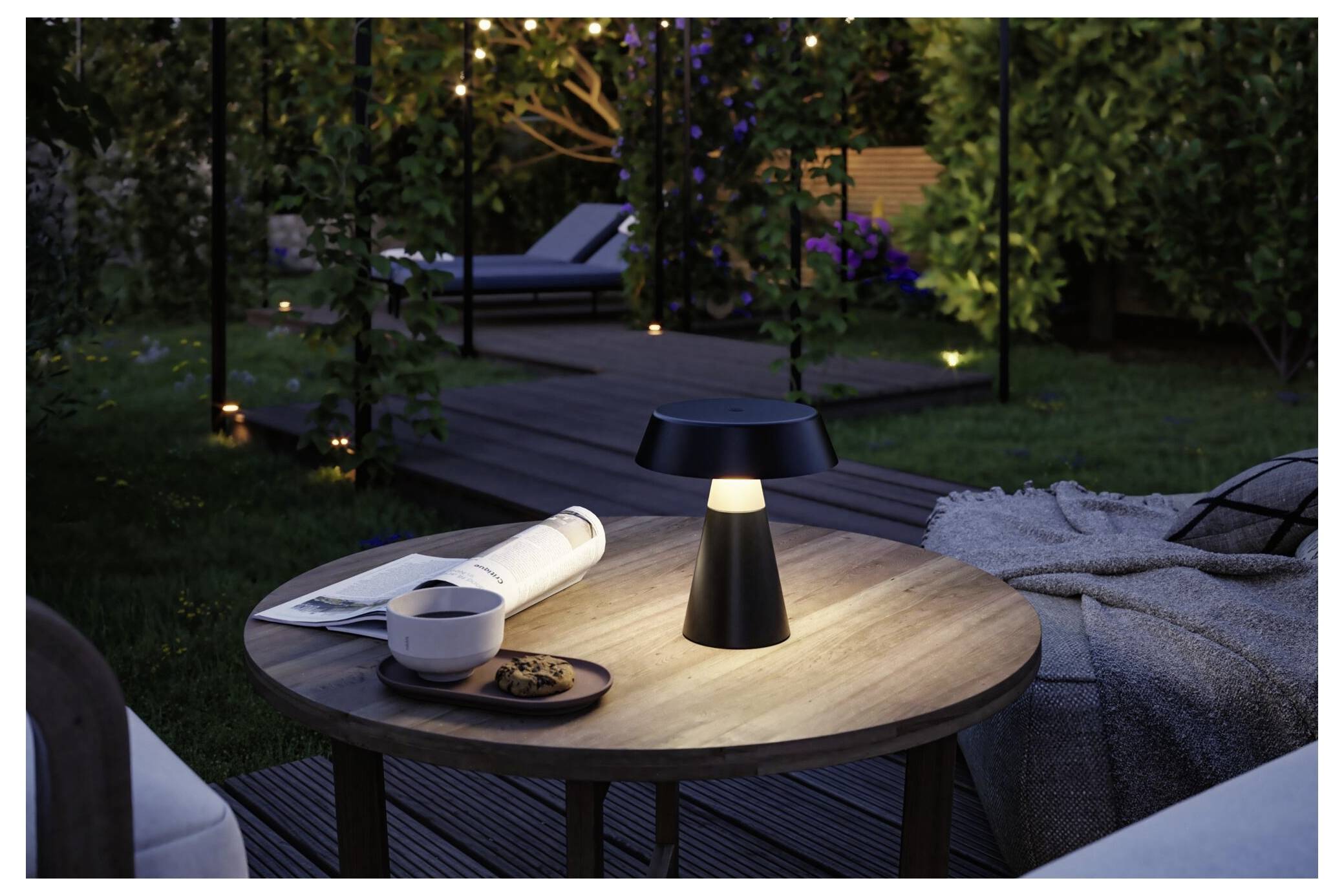 Ein beleuchteter Outdoor-Patiottisch mit einer modernen Lampe, einer Tasse, einer Zeitschrift und Keksen neben einem gepolsterten Loungesessel in der Dämmerung.