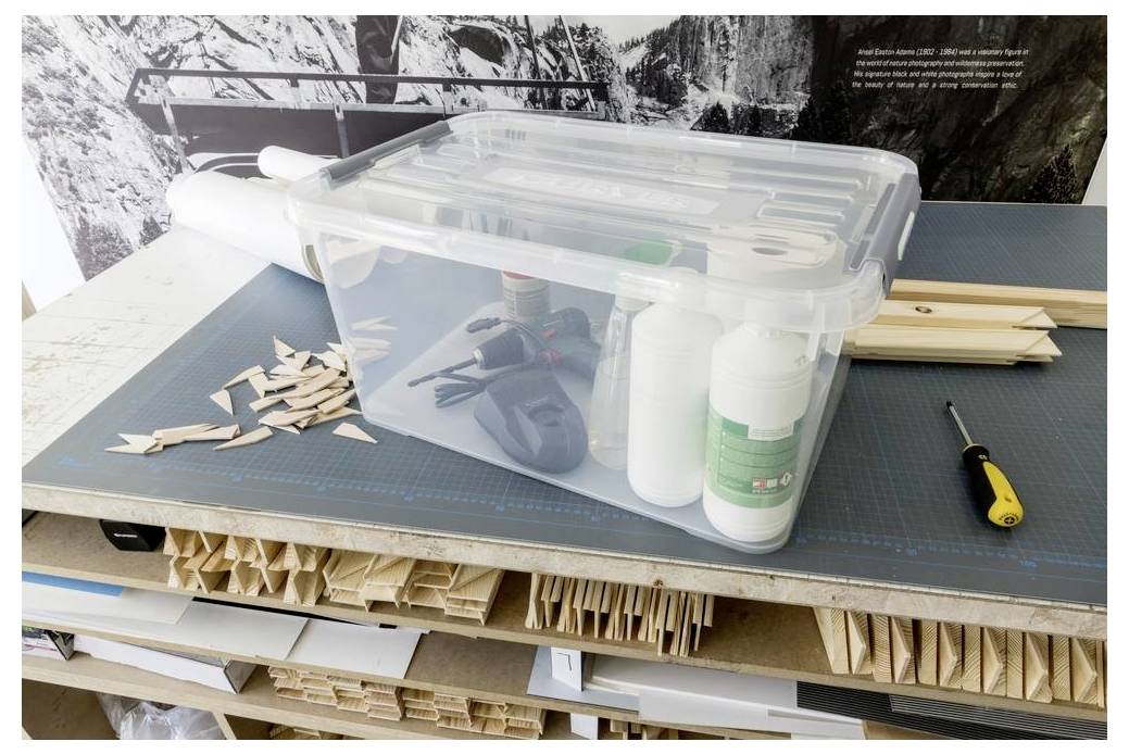 Kunststofflagerbehälter, der eine Werkbank mit Bastelmaterialien, Werkzeugen und geschnittenen Holzstücken abdeckt, was auf einen organisierten Arbeitsbereich oder ein Modellbauprojekt hindeutet.
