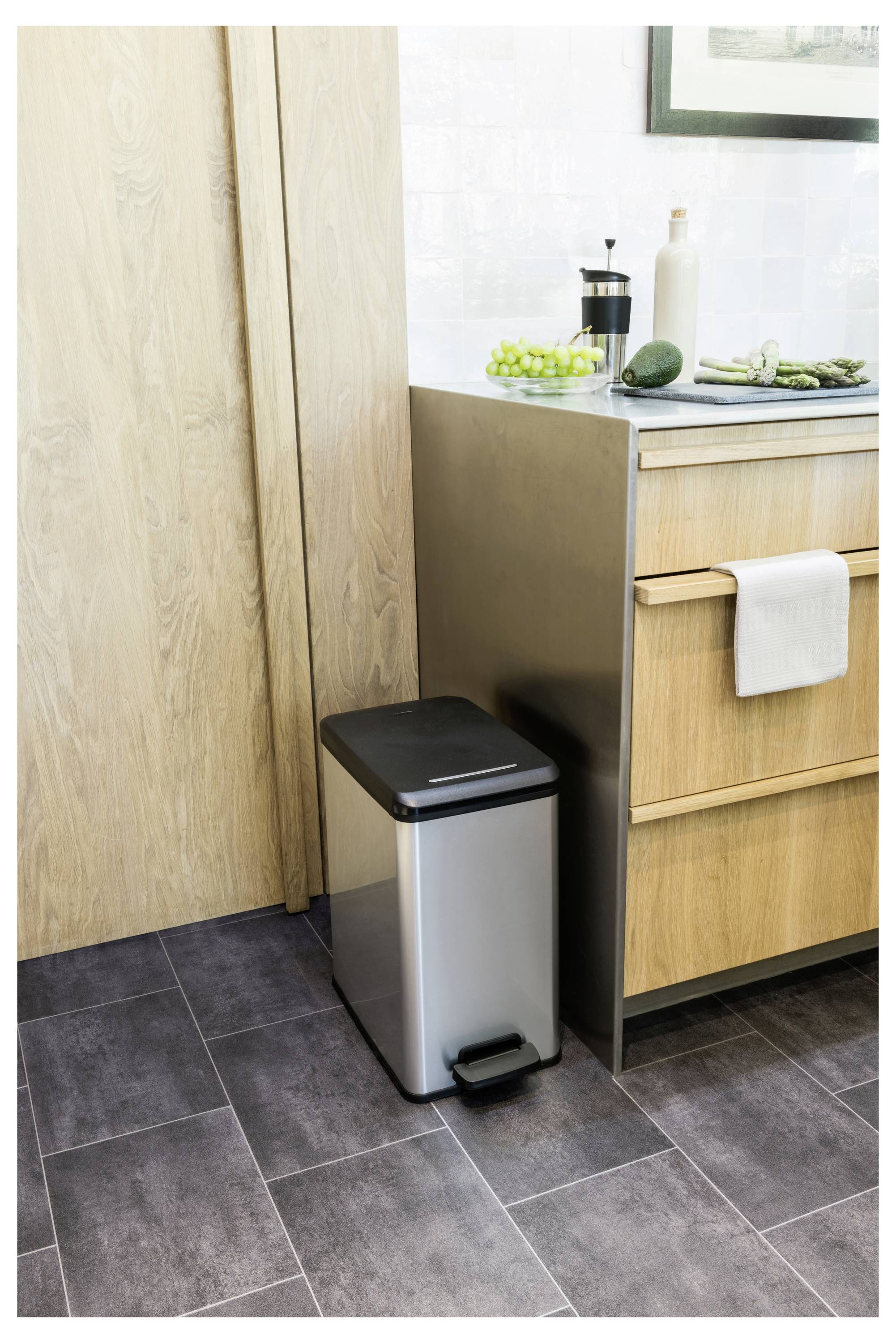 Küchenecke mit einem Edelstahl-Mülleimer neben Holzschränken und einer Arbeitsplatte, auf der eine Kaffeepresse, Trauben und Gemüse stehen.