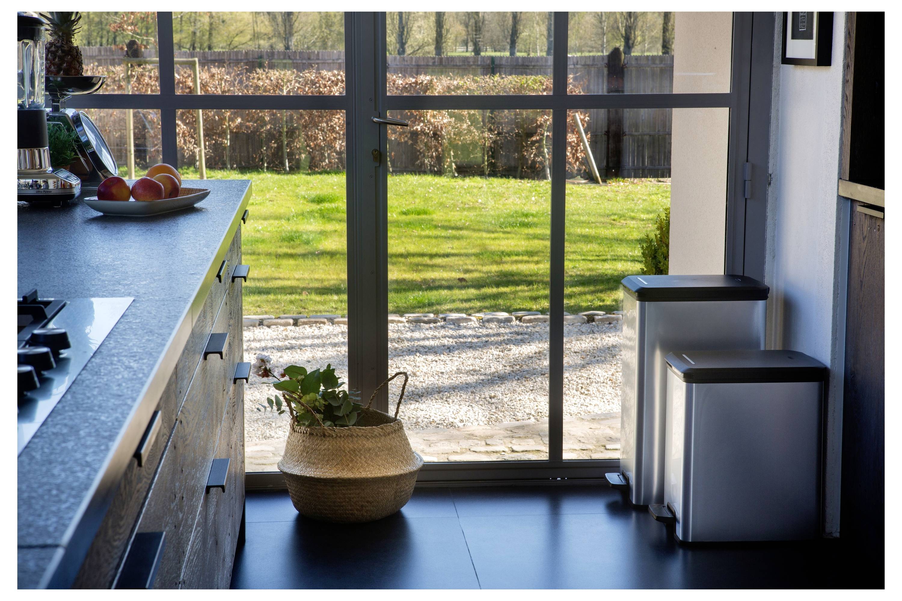 Sonnendurchflutete Küche mit Insel, Obstschale, Topfpflanze und zwei Mülleimern neben Glastüren mit Blick auf einen grünen Garten.