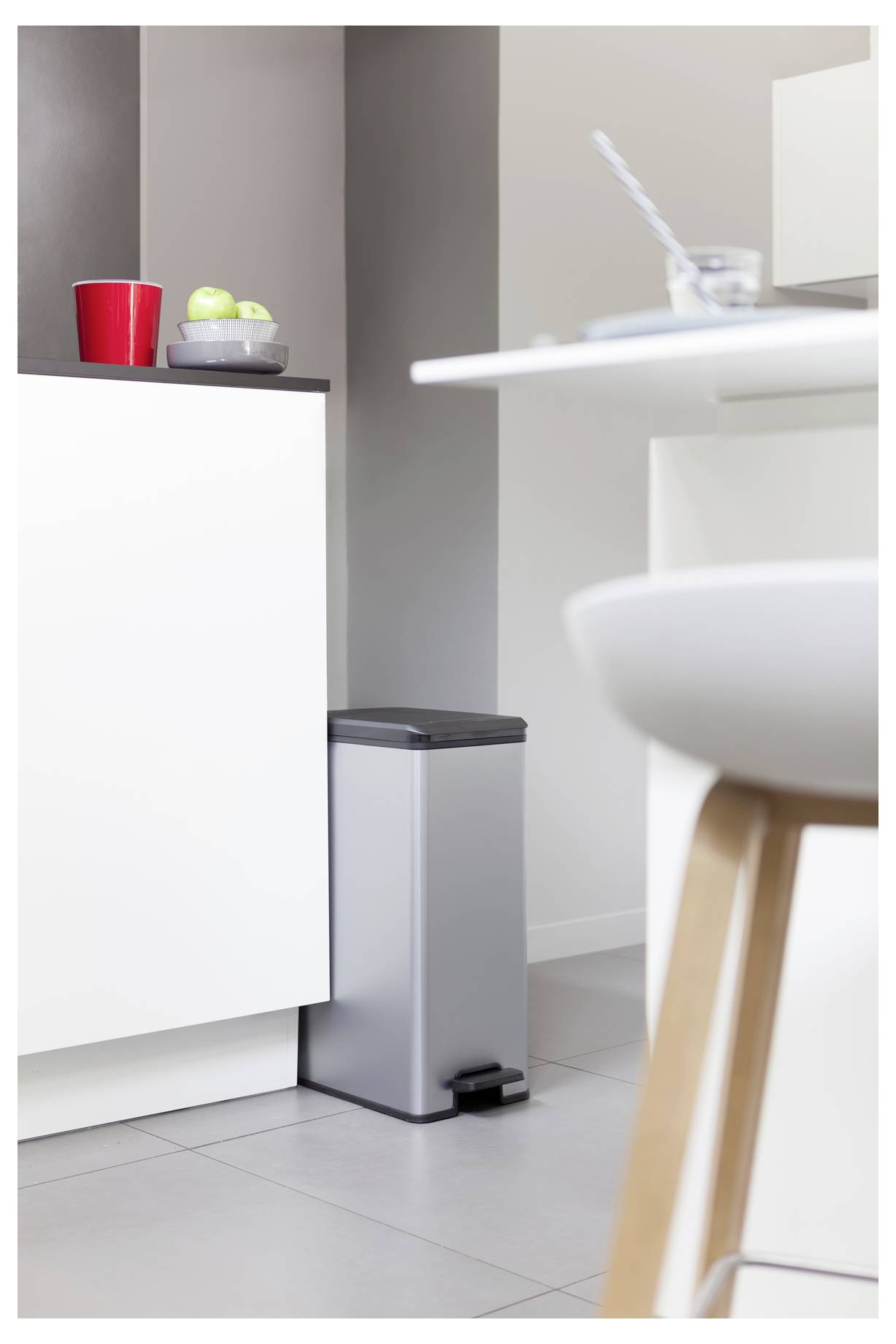 Moderne Kückenecke mit einem silbernen Trittabfalleimer neben einem weißen Schrank, einer roten Tasse und grünen Äpfeln auf der Arbeitsplatte, und einem unscharfen Esszimmerstuhl im Vordergrund.