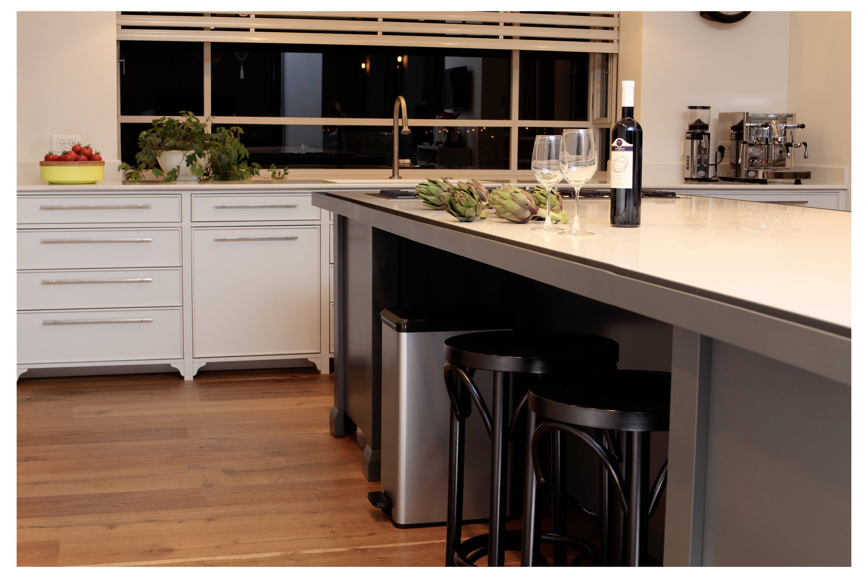 Moderne Küche mit großer Insel, Weinflasche und Gläsern, Artischocken, Barhockern, Spüle, Schränken und Fenster über der Arbeitsplatte.