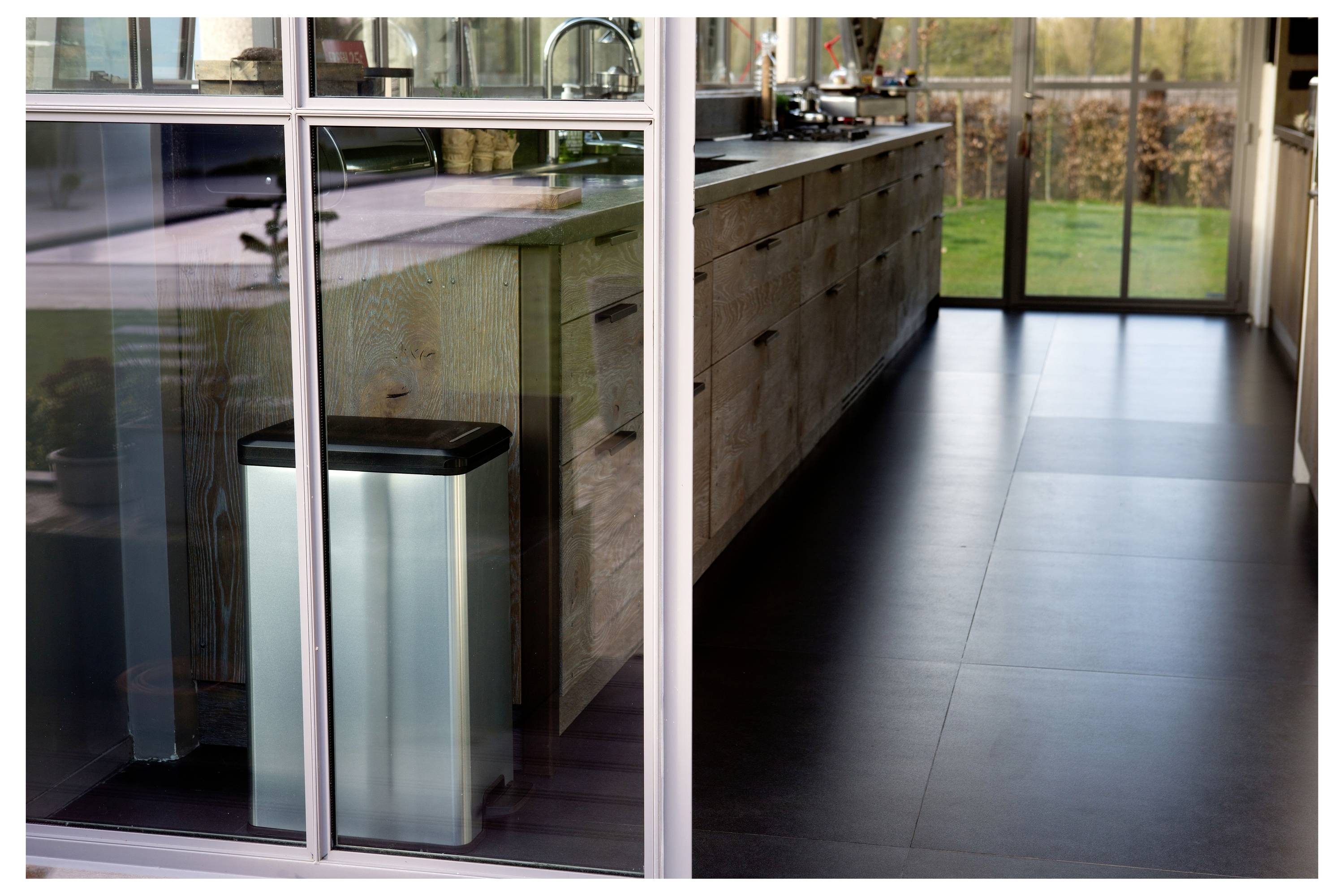 Moderner Flur mit Glastrennwänden, dunklem Fliesenboden und einem beleuchteten Mülleimer in der Nähe einer Rezeptionstheke oder Küchenzeile.