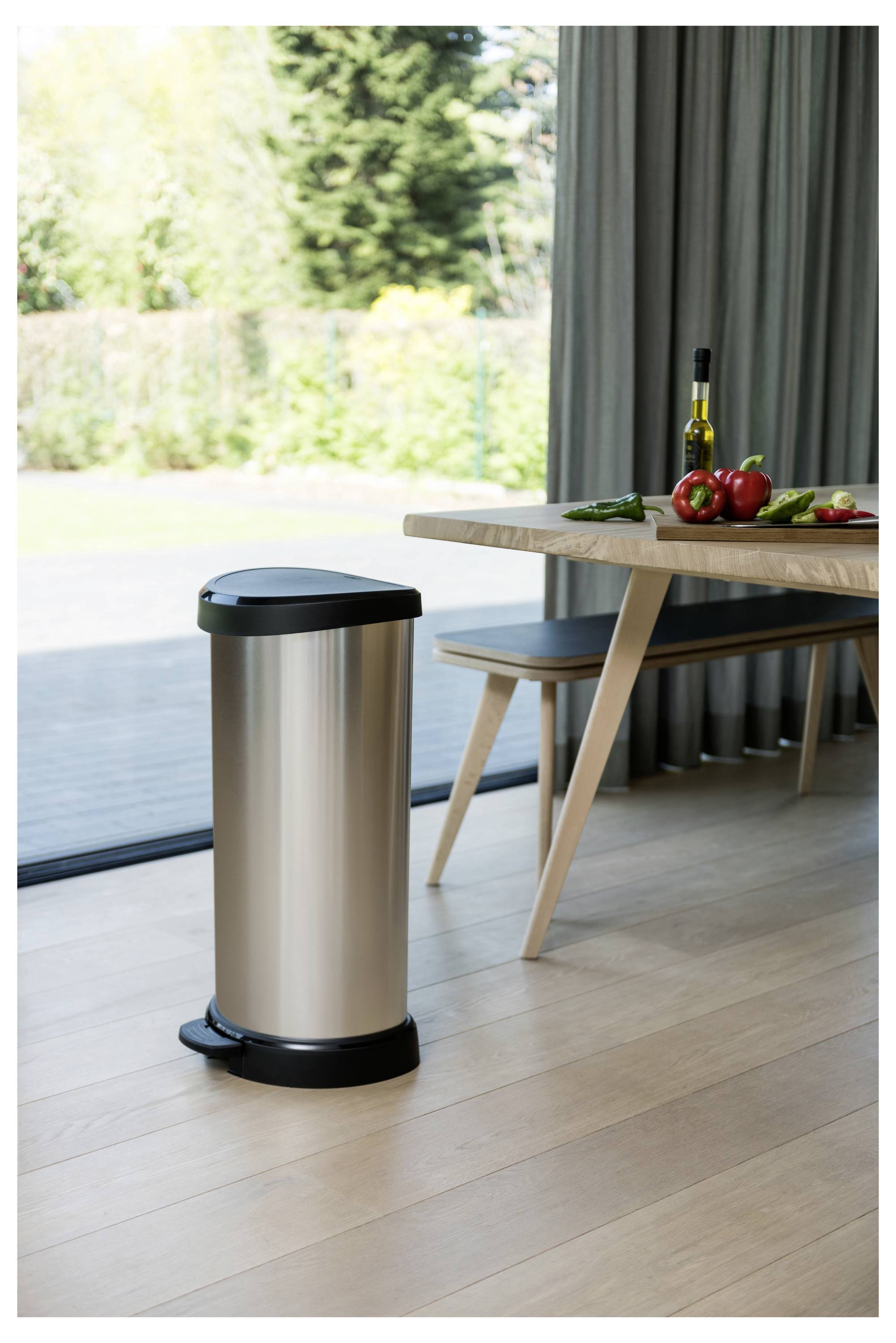 Edelstahlmülleimer in der Küche auf einem Holzboden neben einem gedeckten Esstisch mit Gemüse und einer Flasche, in der Nähe einer Schiebeglasstür.