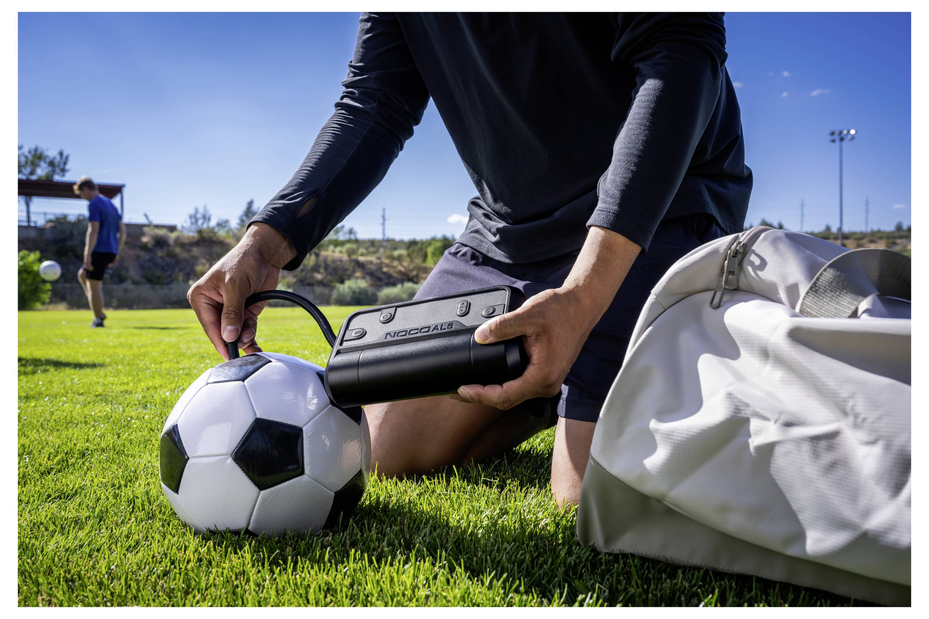 Fußballspieler bläst einen Ball auf dem Gras neben einer Sporttasche auf, während ein anderer Spieler im Hintergrund trainiert.