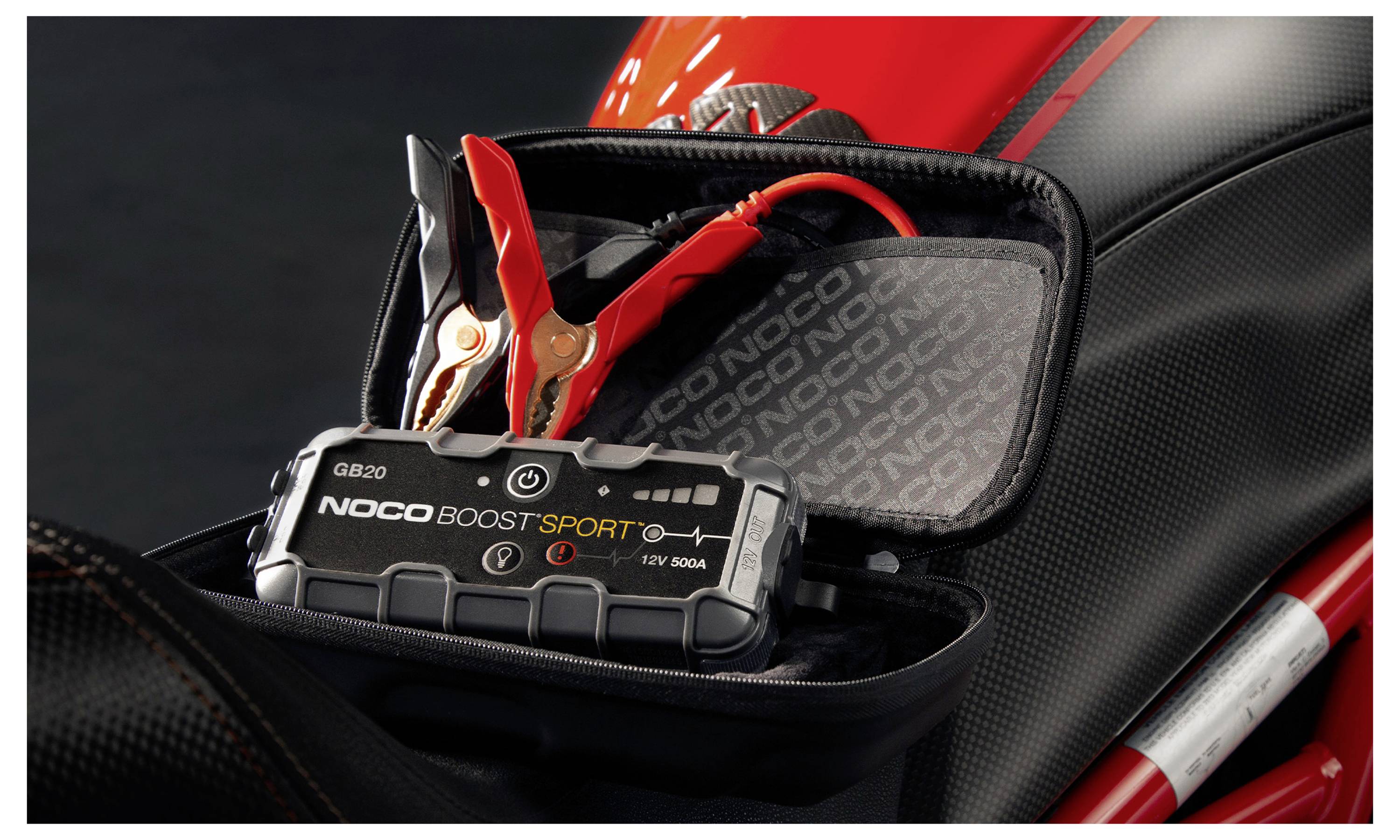 Ein tragbarer NOCO Boost Sport Starthilfegerät mit roten Klemmen sitzt in einem offenen Reißverschluss-Koffer auf einem rot-schwarzen Fahrzeugsitz.