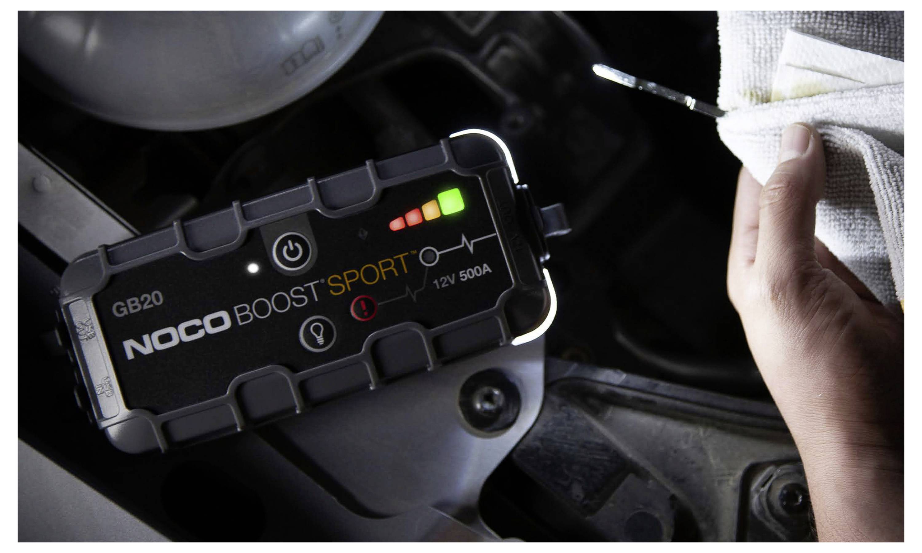 Tragbarer NOCO Boost Sport Starthilfegerät, das in einem Motorraum eines Autos angeschlossen ist, mit einer Hand, die in der Nähe Starthilfekabel oder Kabel hält.