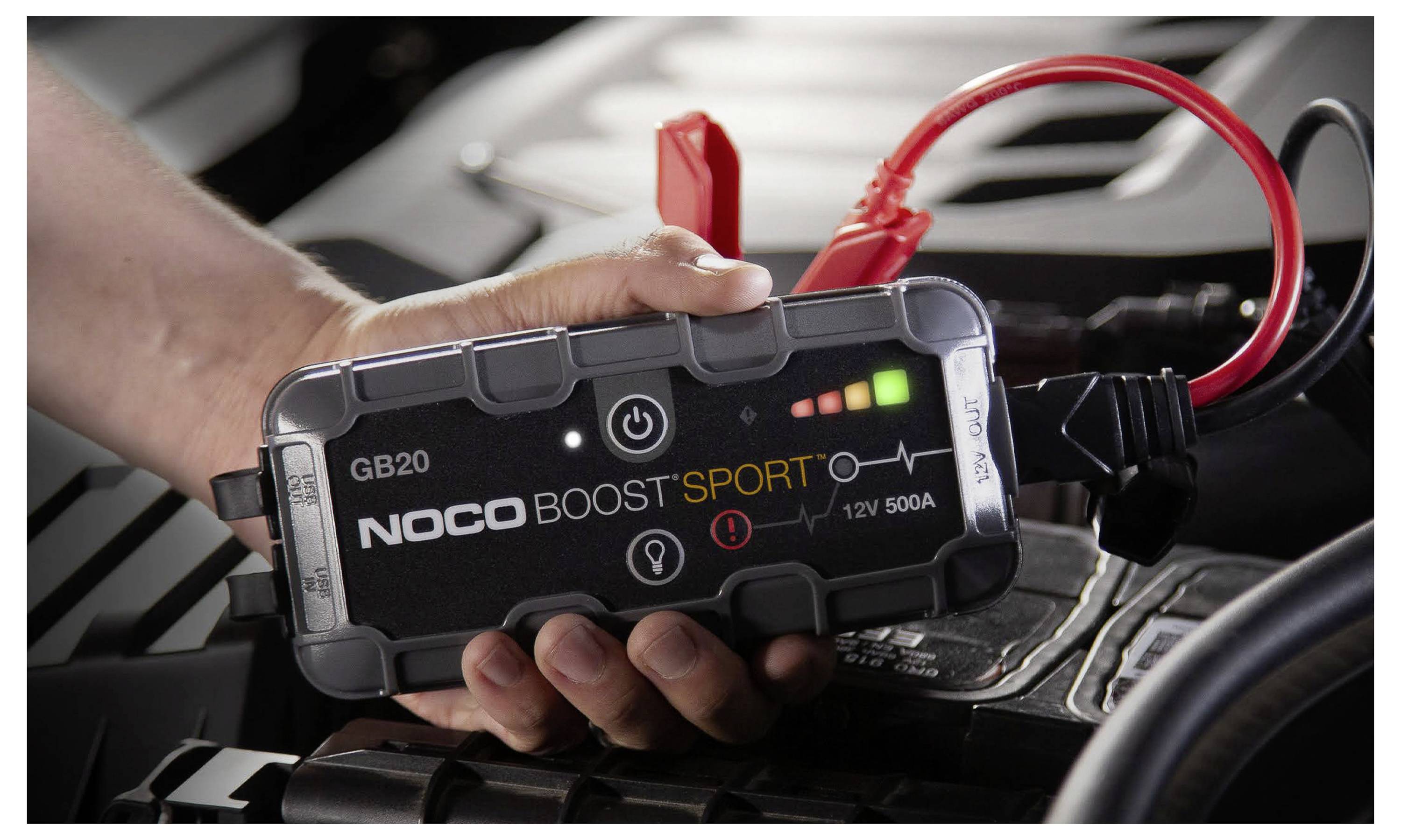 Hand hält einen NOCO Boost Sport tragbaren Autostarthilfegerät, das mit roten Kabeln an einer Fahrzeugbatterie angeschlossen ist.